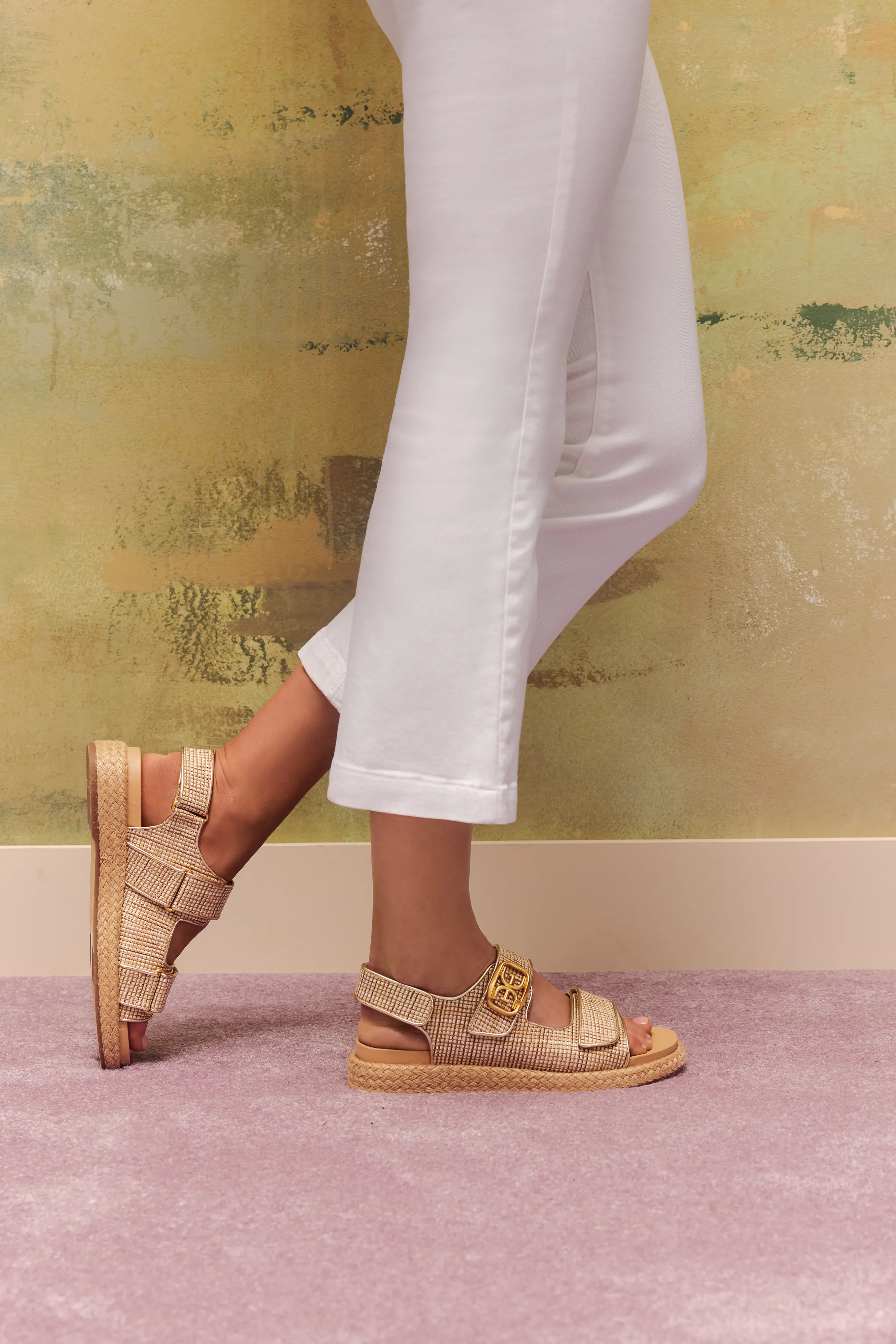 Model in cropped white trousers wearing tan woven strap sandals with gold buckle against a painted green wall, for Sam Edelman.