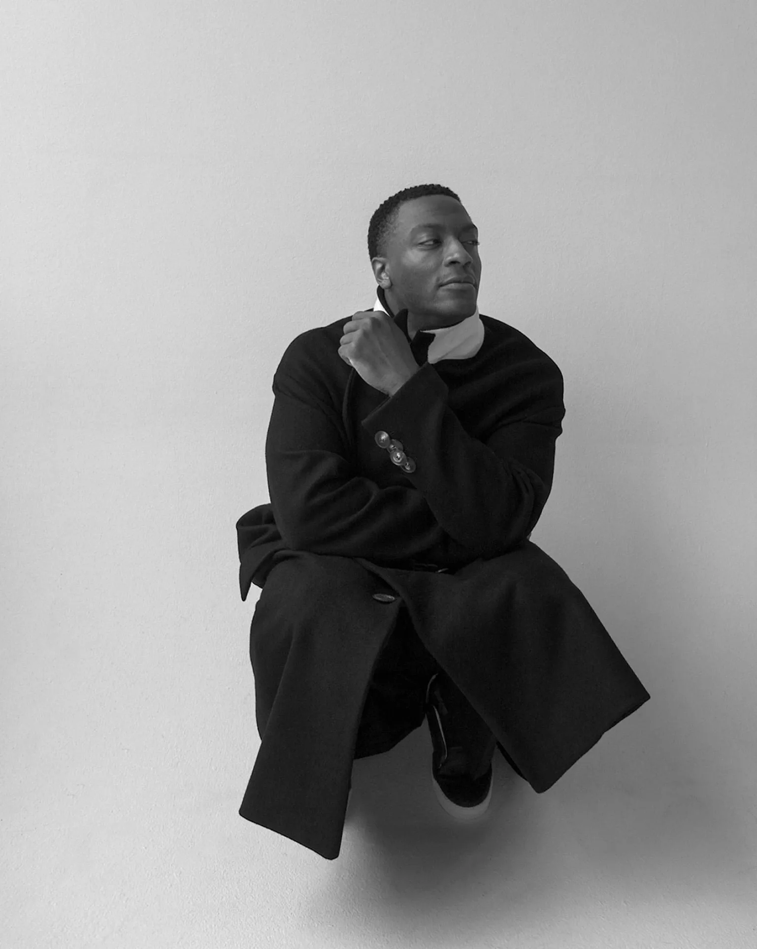 Black and white portrait of a man crouching in a long black overcoat against a plain grey wall, for The Last Magazine.