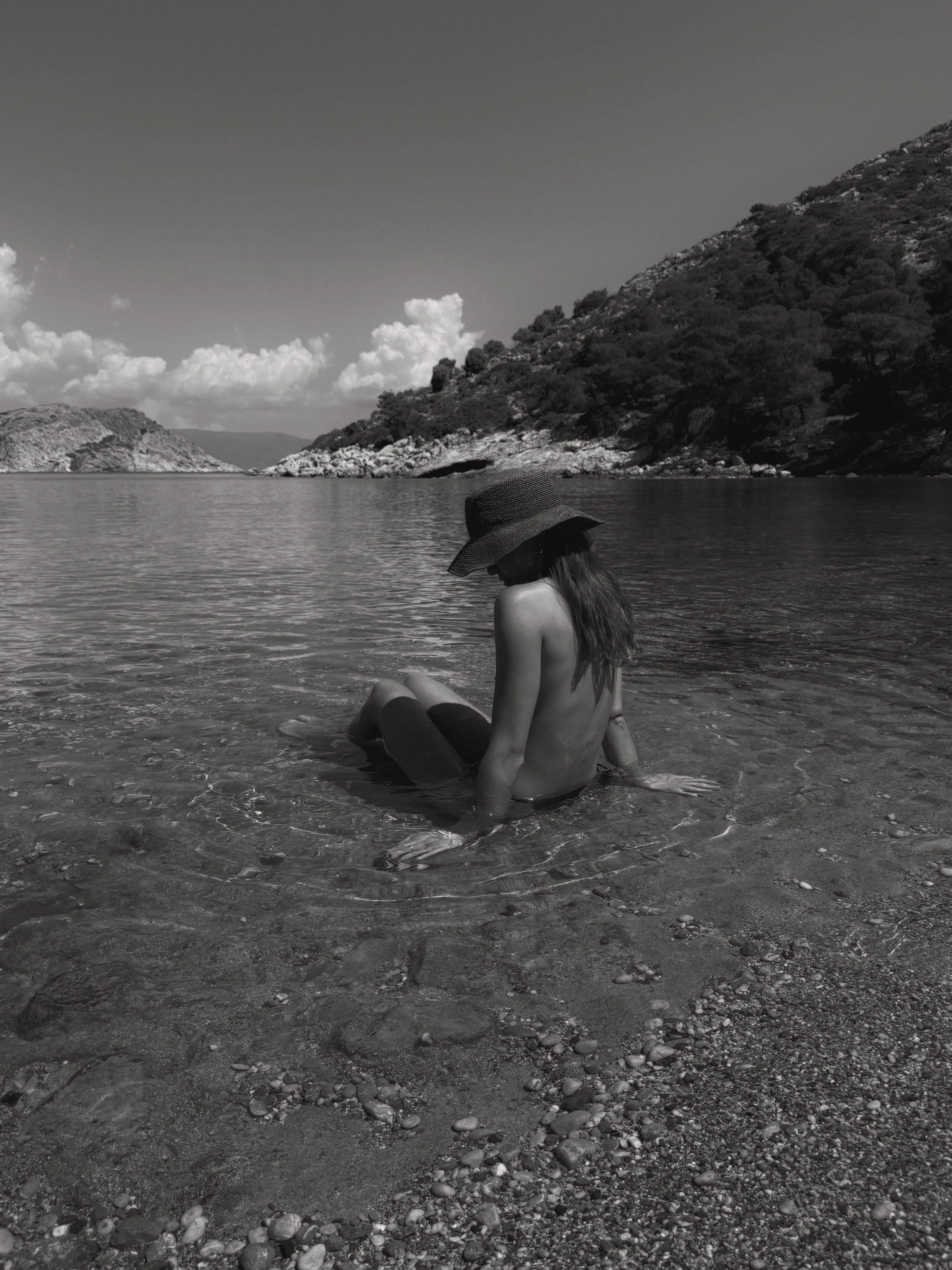 Black-and-white shot of a woman in a wide-brim hat sitting in shallow clear water on a pebble beach by a wooded cove.