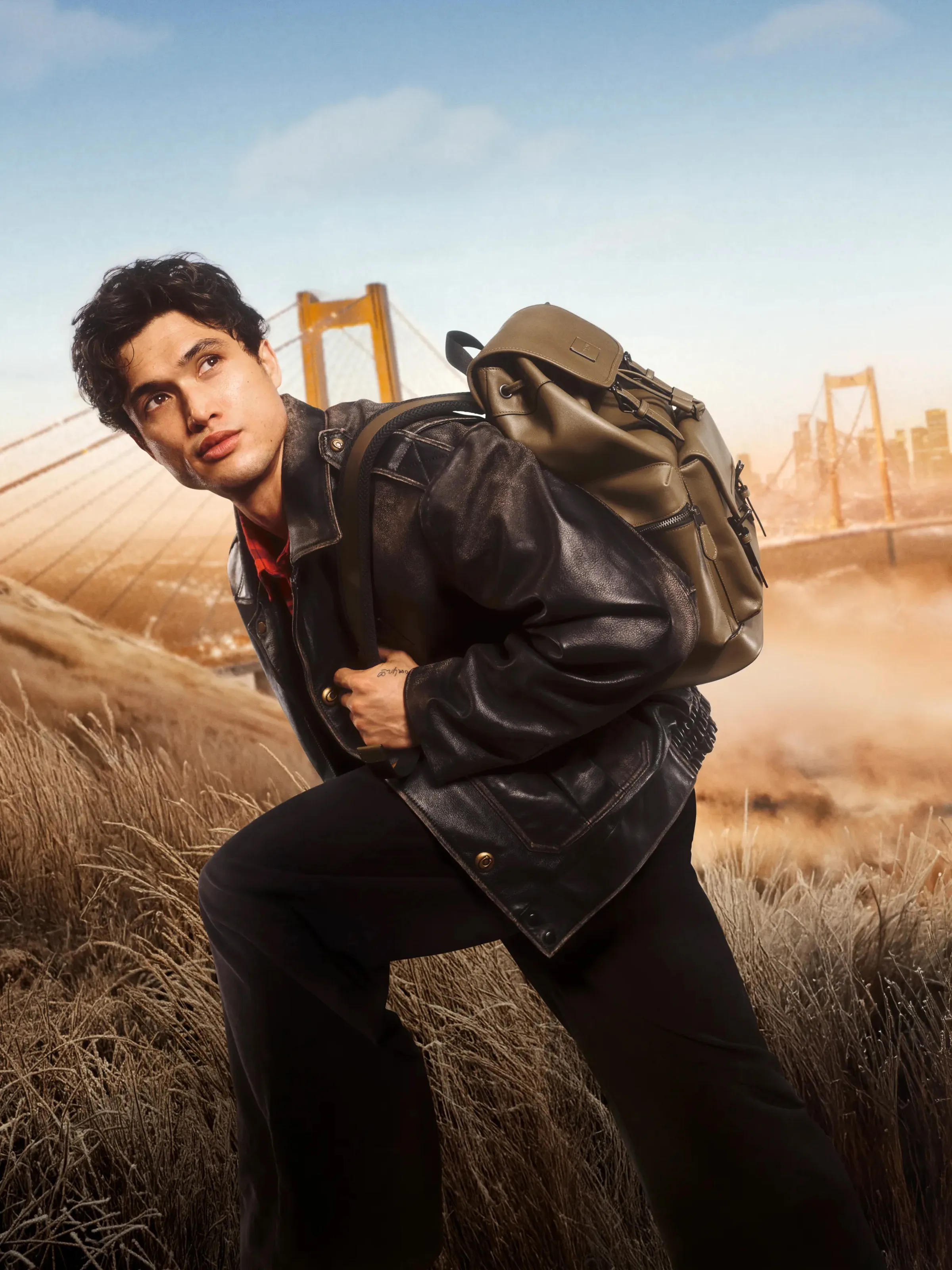 AI image of a man in a black leather jacket carrying an olive backpack in tall grass near a golden bridge, for Coach.
