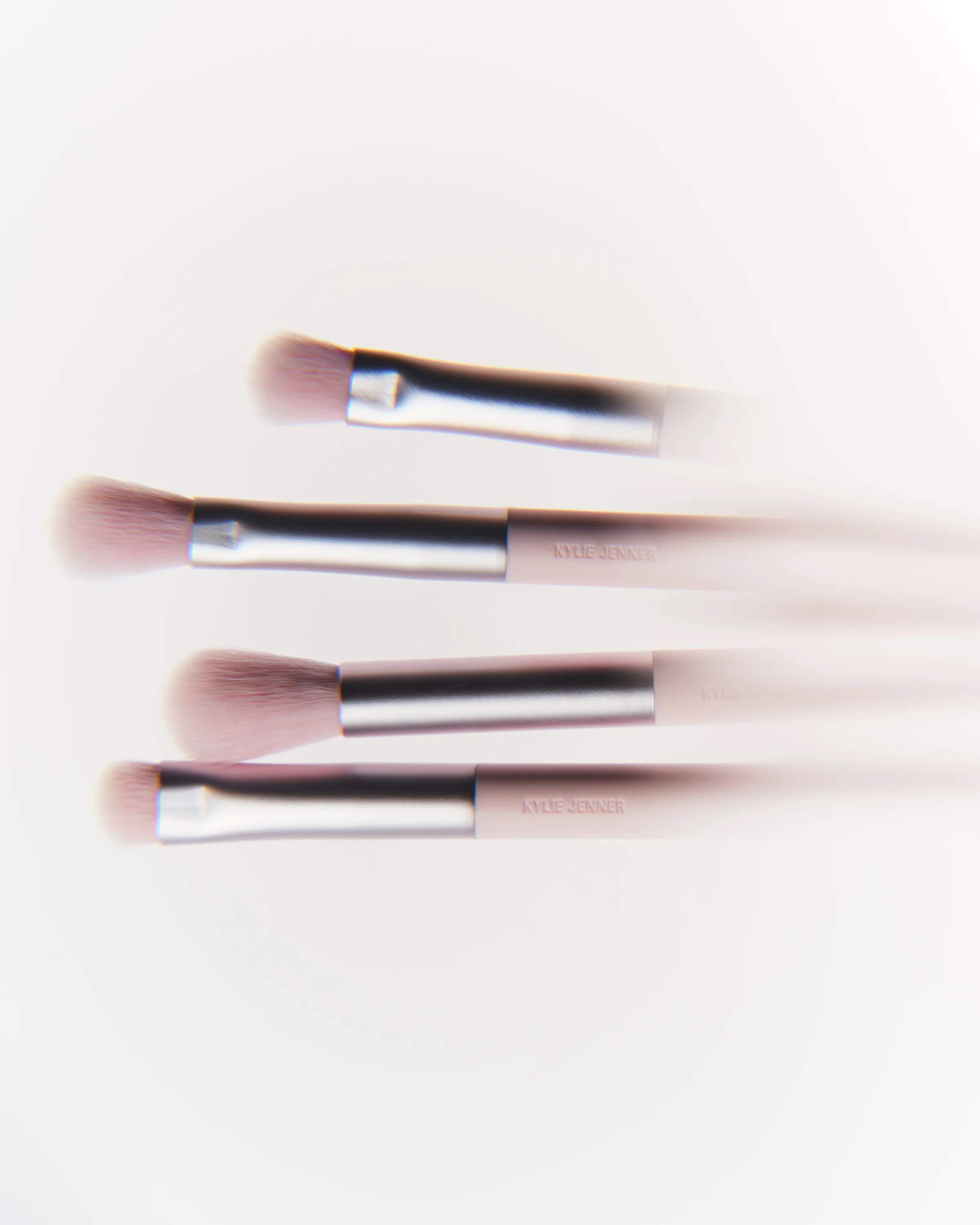 Four pink eyeshadow brushes with silver ferrules lying on white, softly blurred with motion streaks, for Kylie Cosmetics.