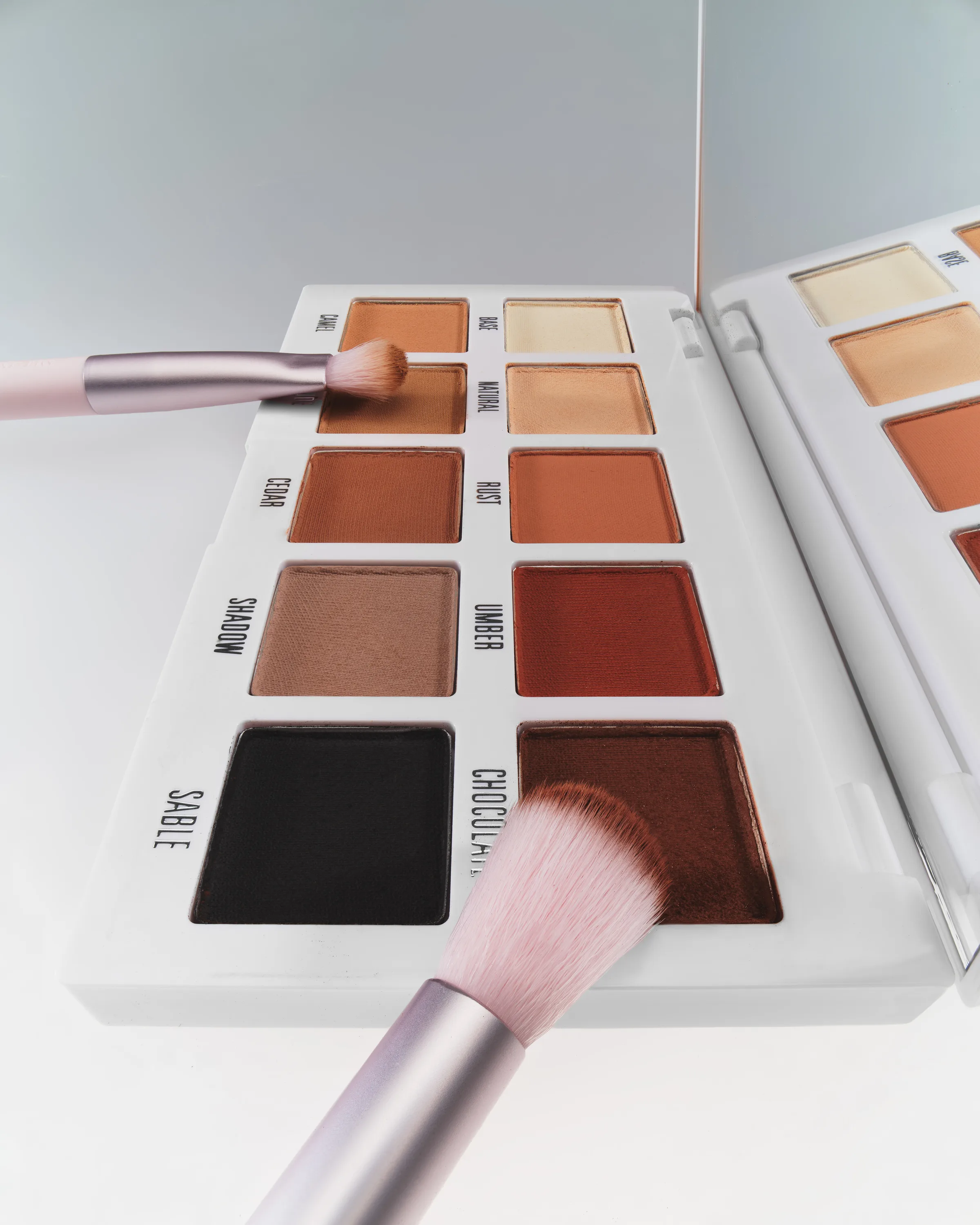 Open matte eyeshadow palette in warm browns with two pink brushes dipping into shades, on white, for Kylie Cosmetics.