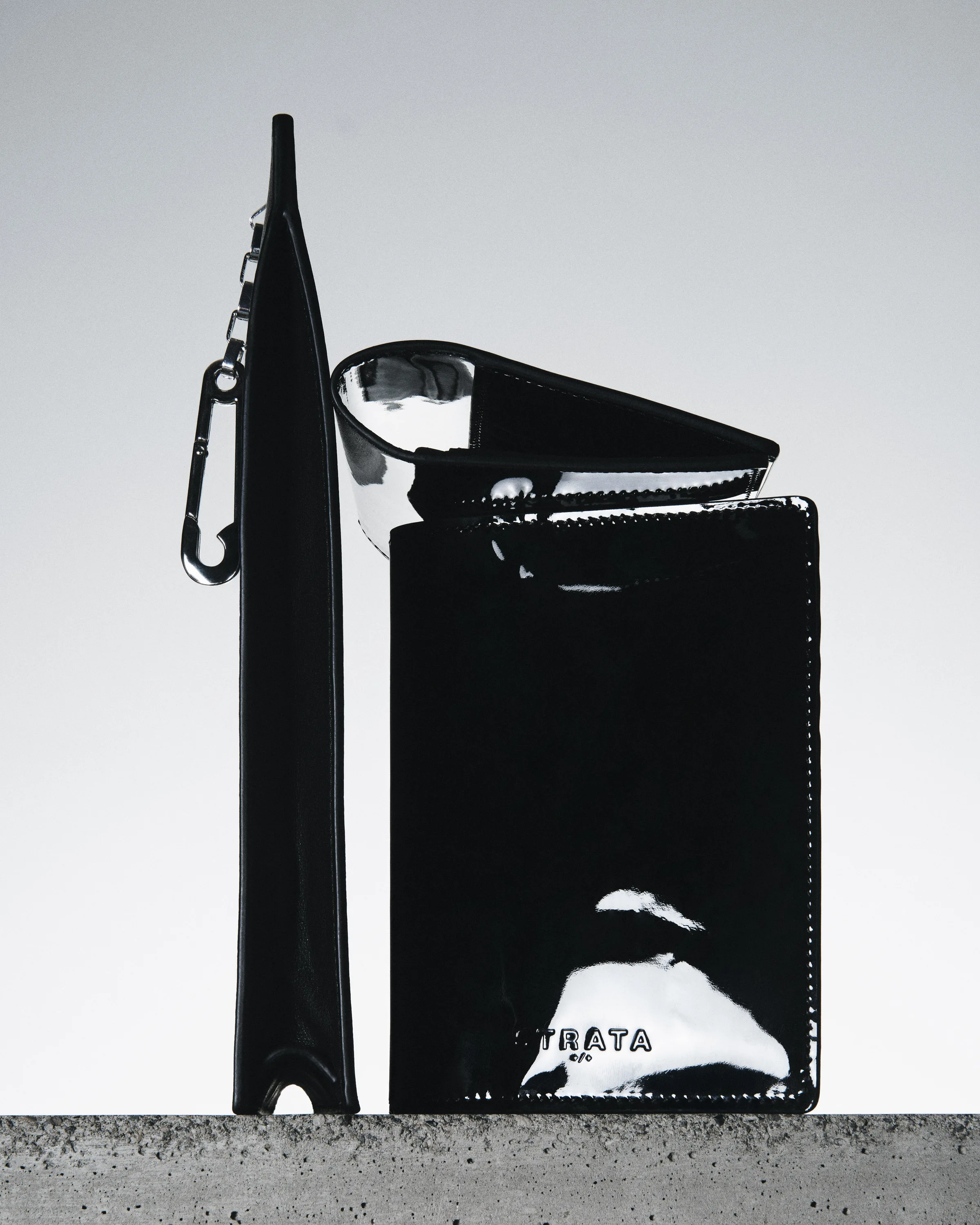 Black patent leather travel sleeve, key holder and pouch standing on a concrete ledge against a grey gradient, for Strata.