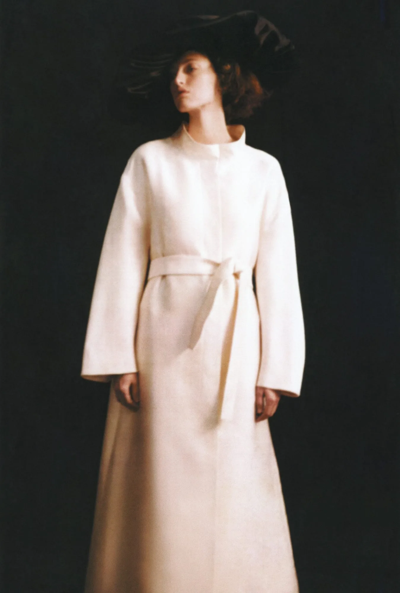 Model in a long white belted coat and wide black hat against a dark backdrop, shot by Amber Witcomb.