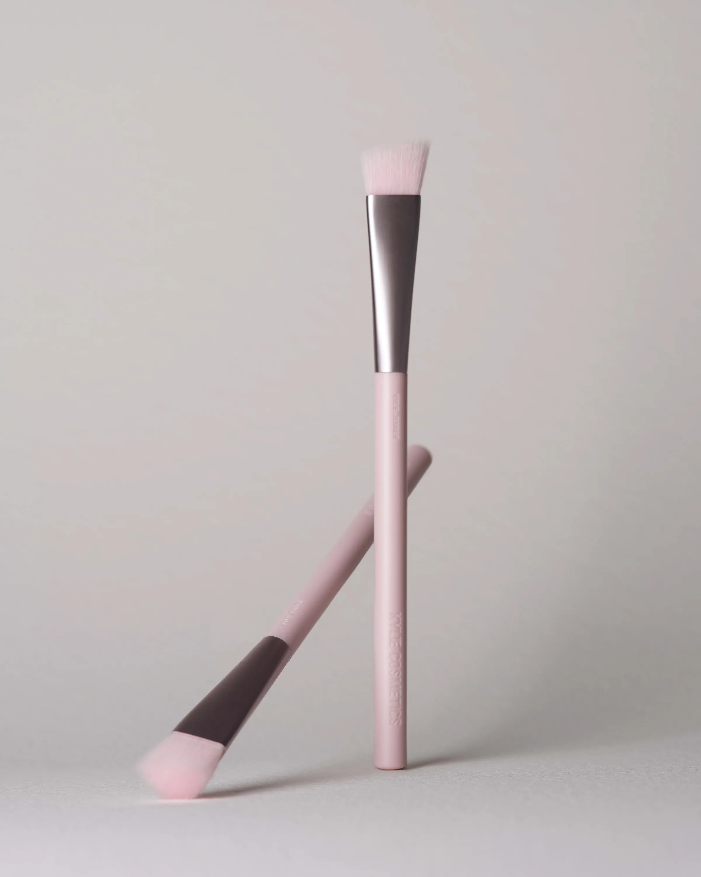 Two pink makeup brushes with angled bristles, one standing upright and one leaning, on a grey backdrop, for Kylie Cosmetics.