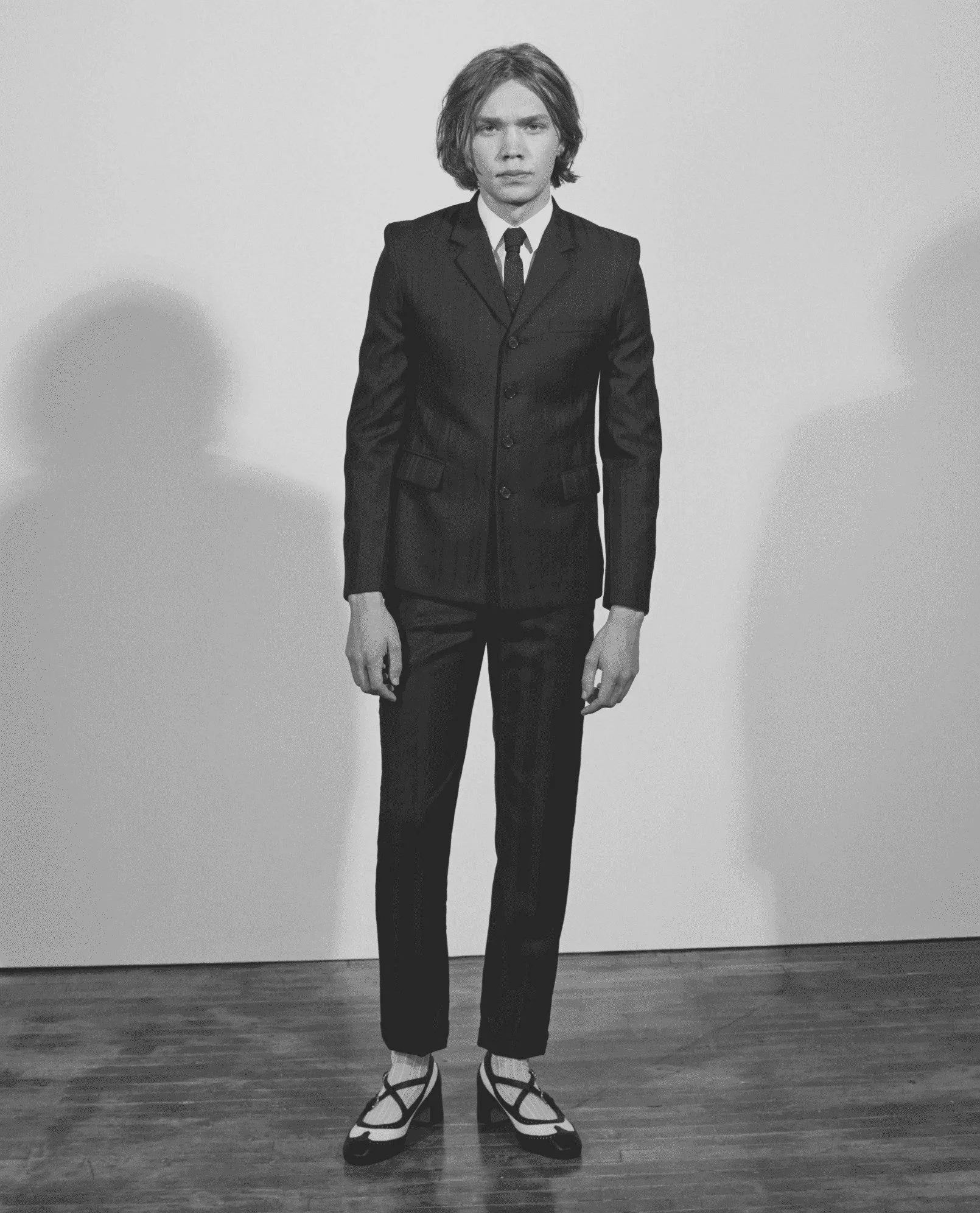 Black and white full-length portrait of a young man in a pinstripe suit and strapped two-tone heels, for L'Officiel Hommes.