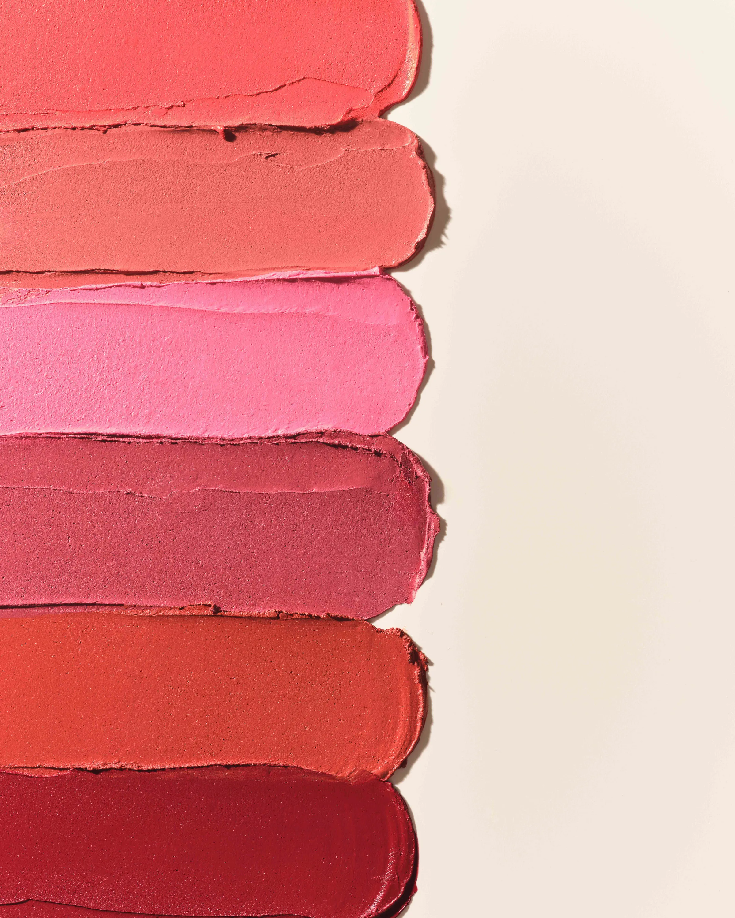 Six lipstick swatches in coral, pink, rose and red smeared in stripes on a cream background, for Kylie Cosmetics.
