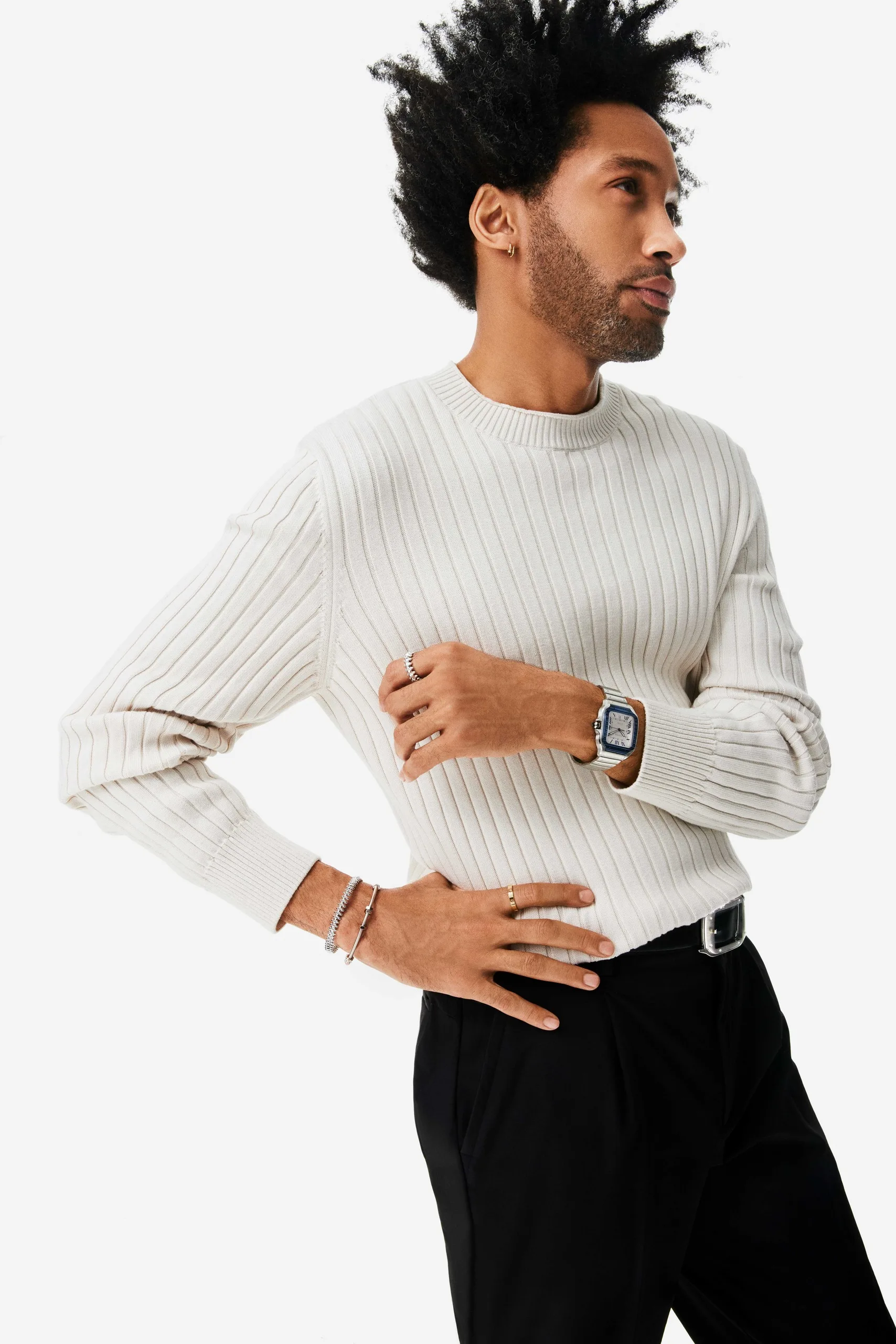 Model in a ribbed cream sweater and black trousers wearing a steel watch, rings and bracelets on white, for Cartier.
