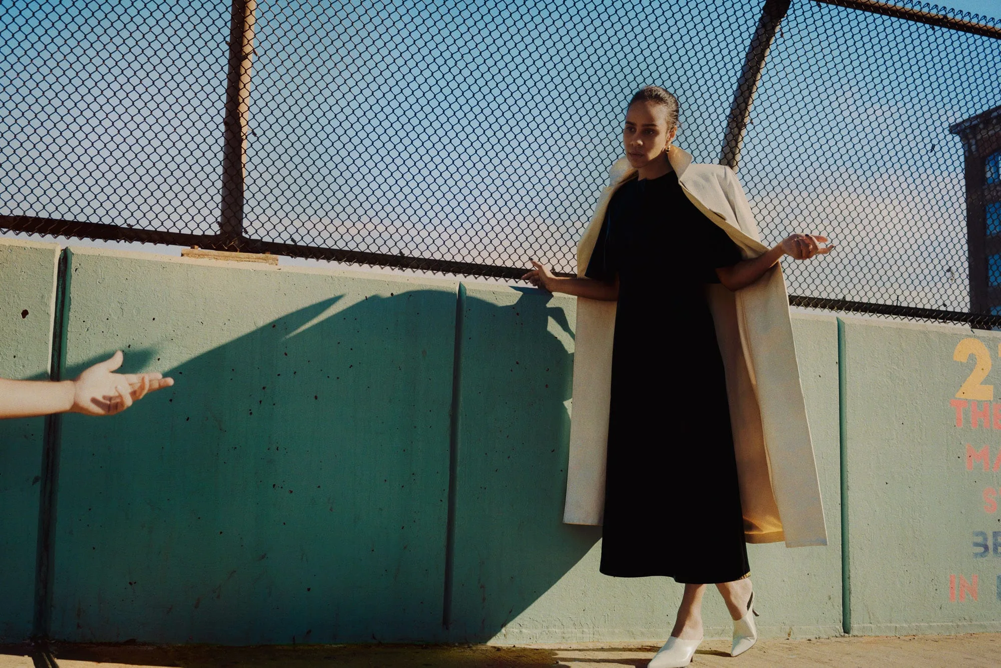 Model in a black dress, cream coat and white heels against a teal wall and chain-link fence, for The Last Magazine.