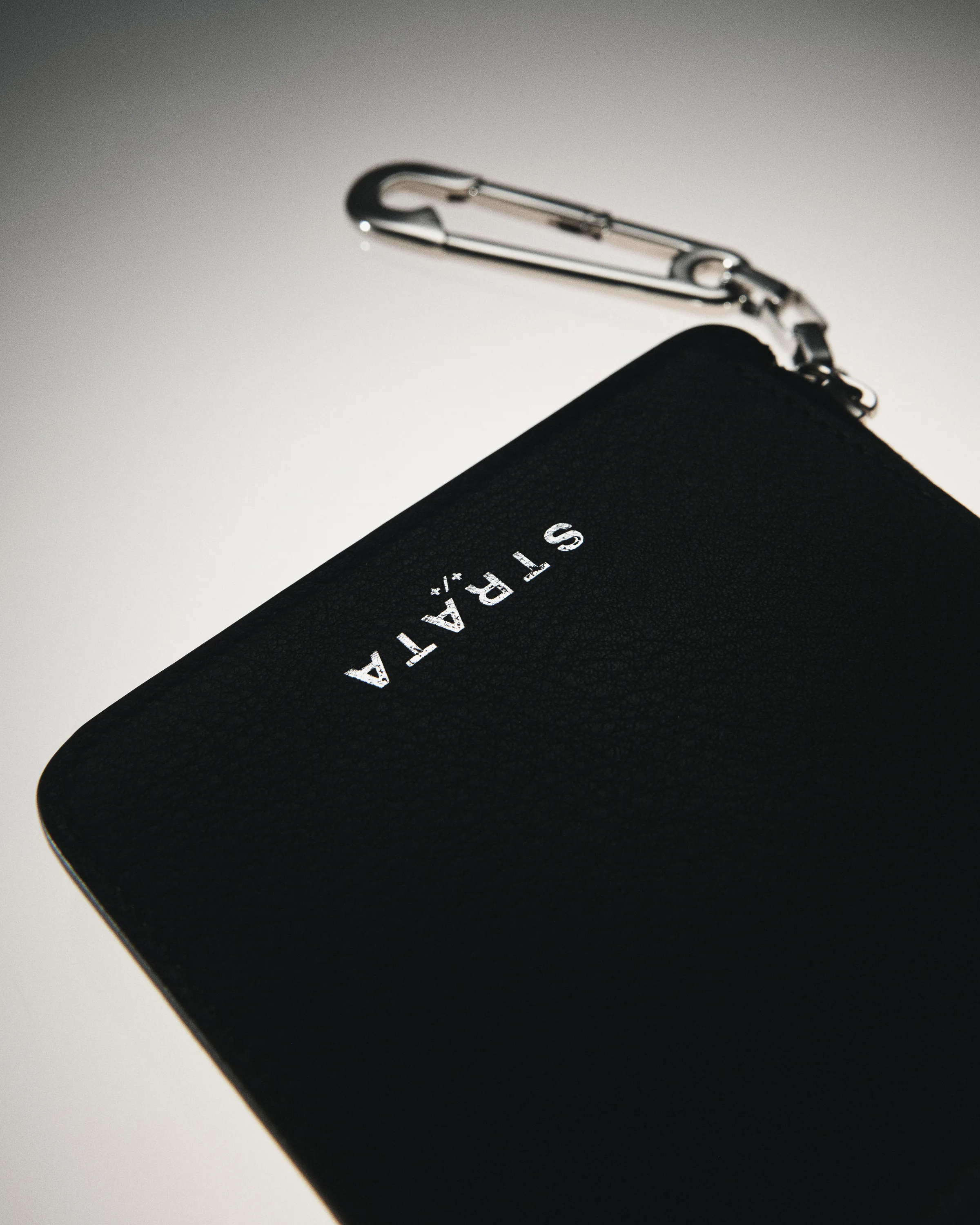 Black pebbled leather zip wallet with silver STRATA logo and safety-pin clasp on a grey gradient, for Strata.