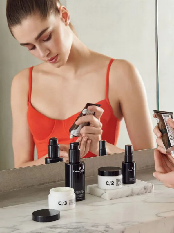 Model in a red camisole applies Costa Brazil skincare at a marble vanity mirror, bottles and jars in front, for Net-a-Porter.