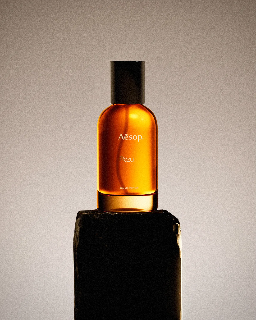 CGI render of an amber Rōzu Eau de Parfum bottle on a dark stone plinth against a soft grey backdrop, for Aesop.