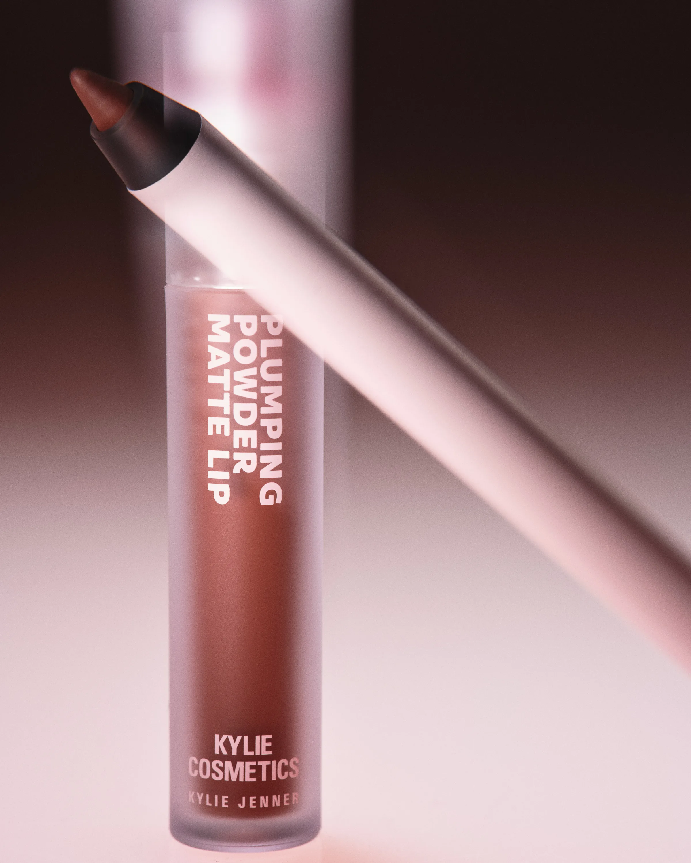 Plumping Powder Matte Lip tube in toffee brown with a white lip liner leaning across it on a pink backdrop, for Kylie Cosmetics.