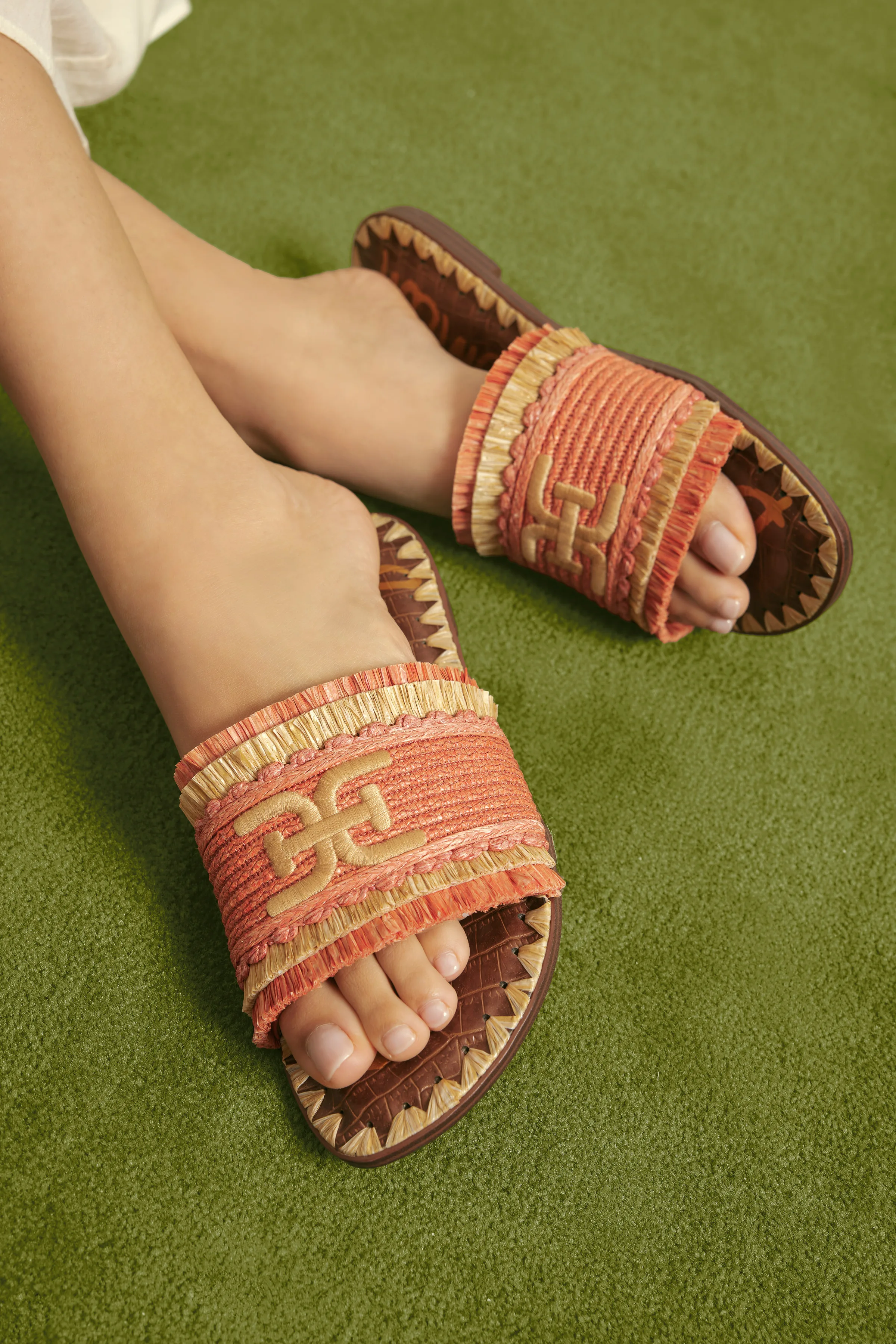 Feet in coral woven raffia slide sandals with gold logo emblem on green carpet, for Sam Edelman.