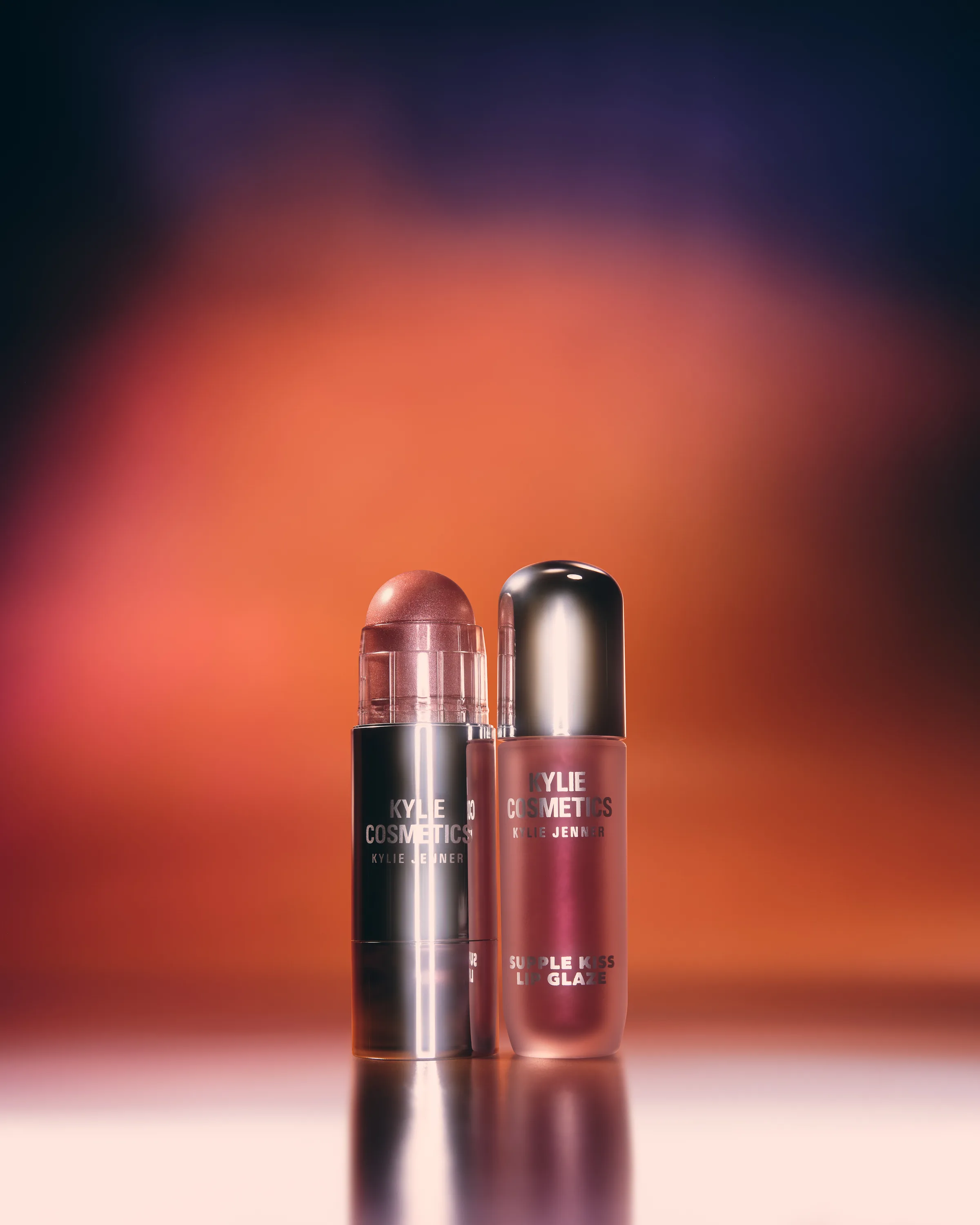Rosy Ray lip glaze and Rose Dusk dewy balm stick on a reflective surface before an orange and purple glow, for Kylie Cosmetics.