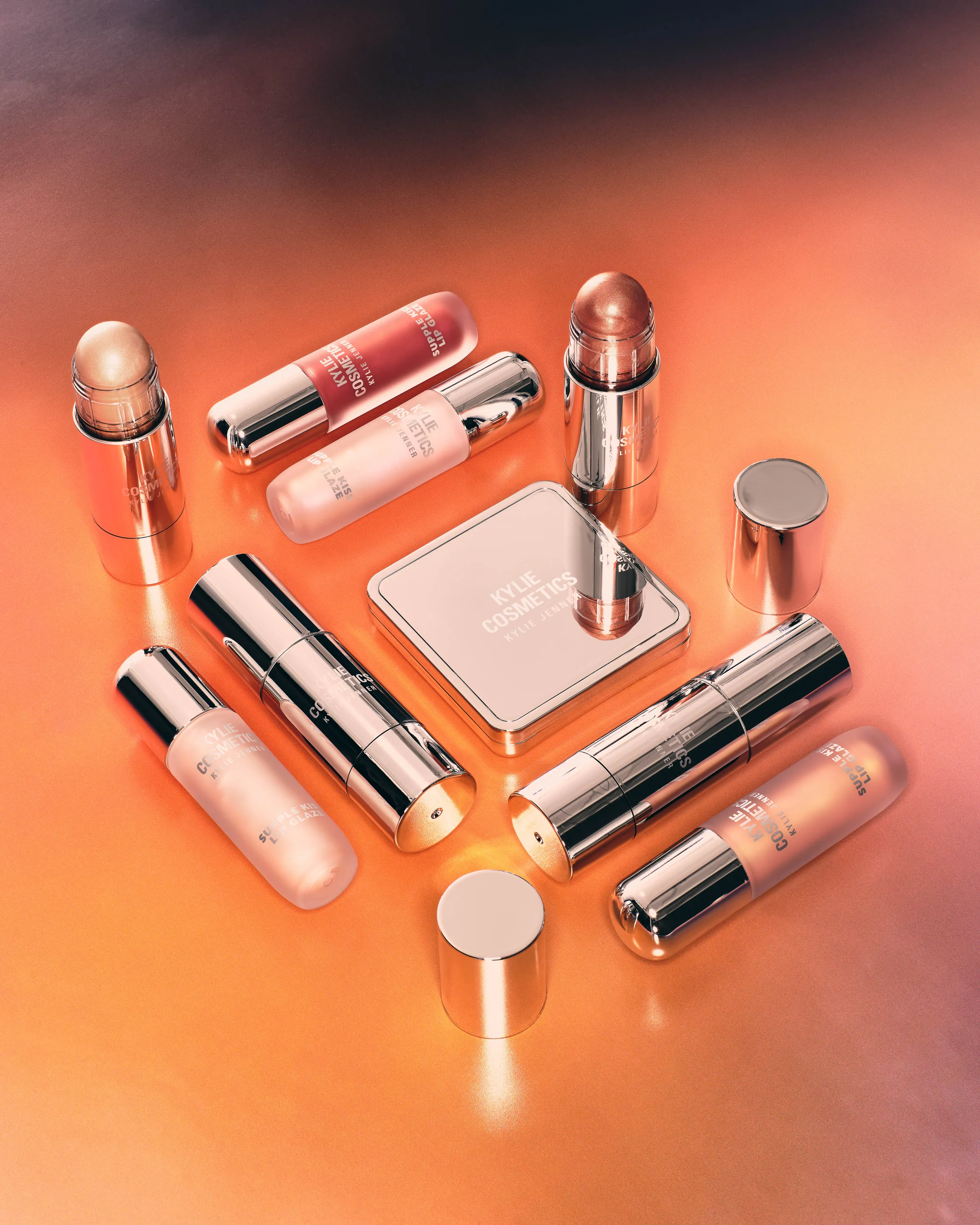 Chrome makeup sticks, glosses and compact arranged on a glowing orange and pink surface, for Kylie Cosmetics.