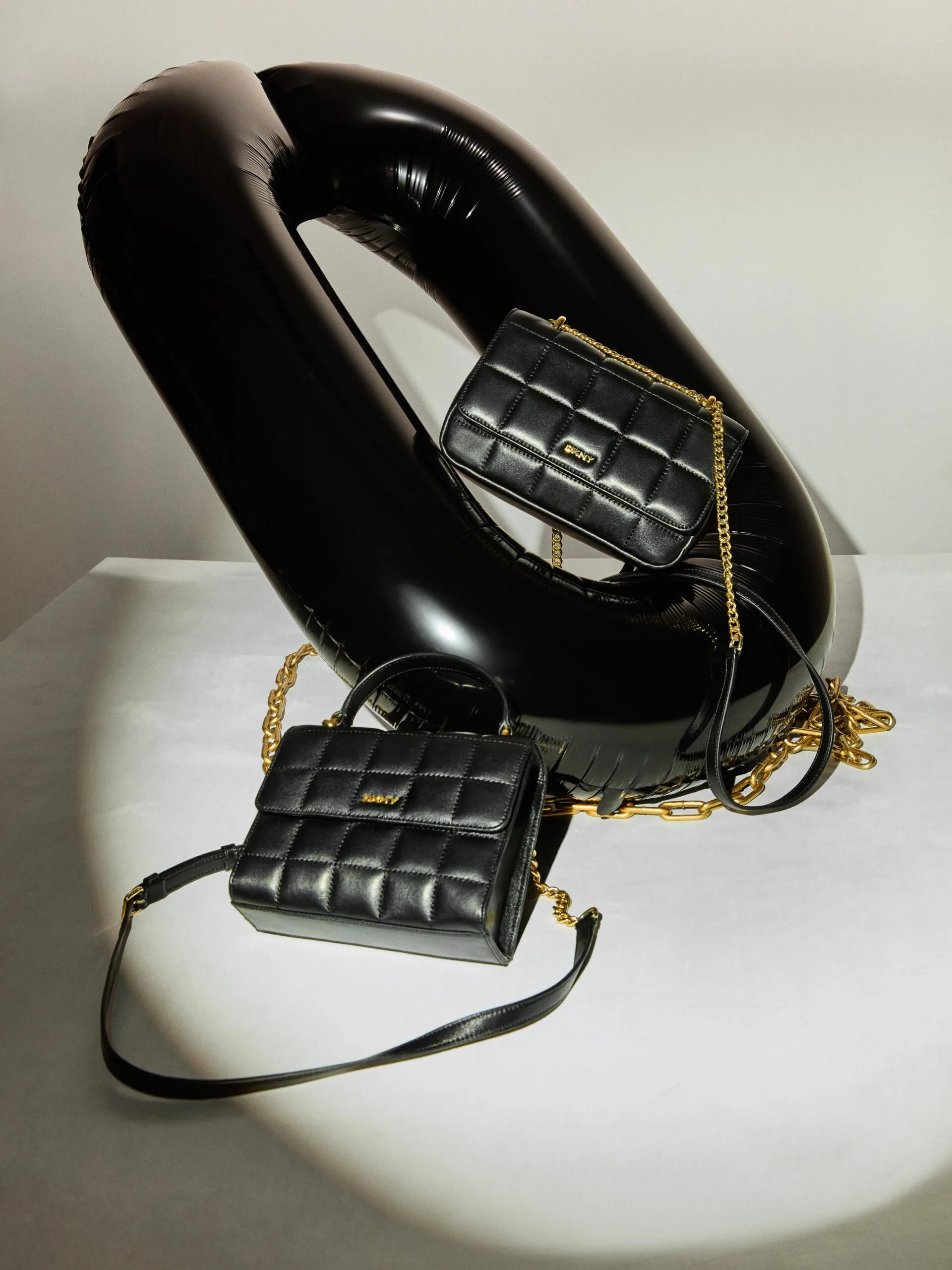 Two black quilted leather bags with gold chain straps posed on a glossy black inflatable loop under a spotlight, for DKNY.