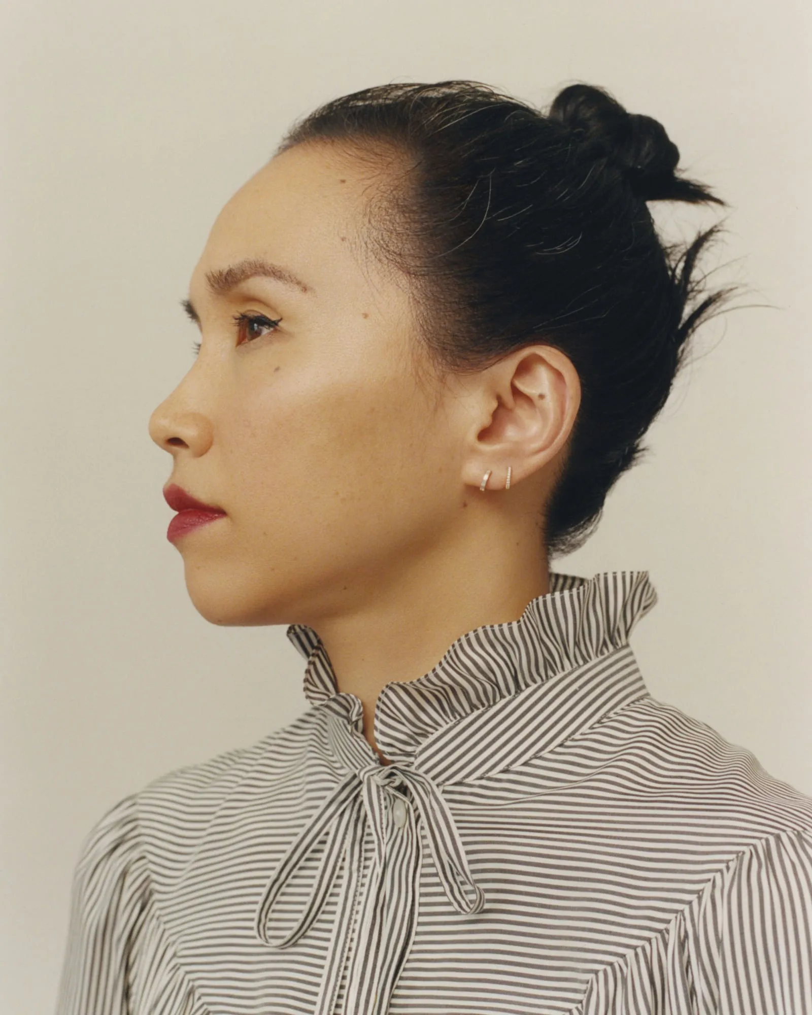Profile portrait of a woman with a top knot, red lips and gold earrings in a striped ruffle-neck blouse, for Vogue Korea.