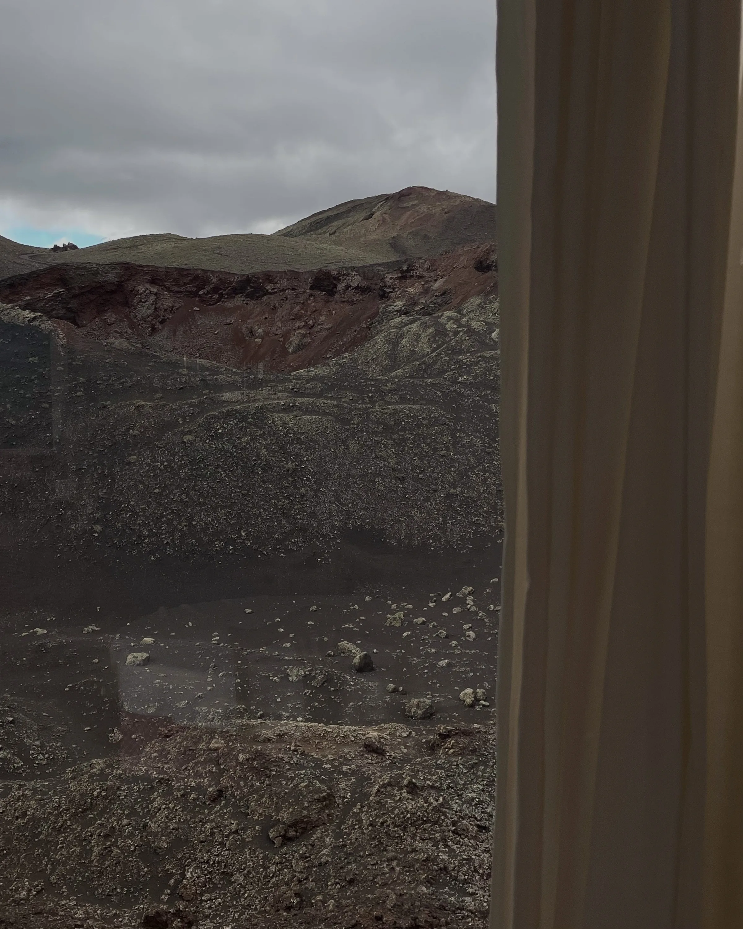 Rocky volcanic crater under grey clouds seen through a window beside a beige curtain.