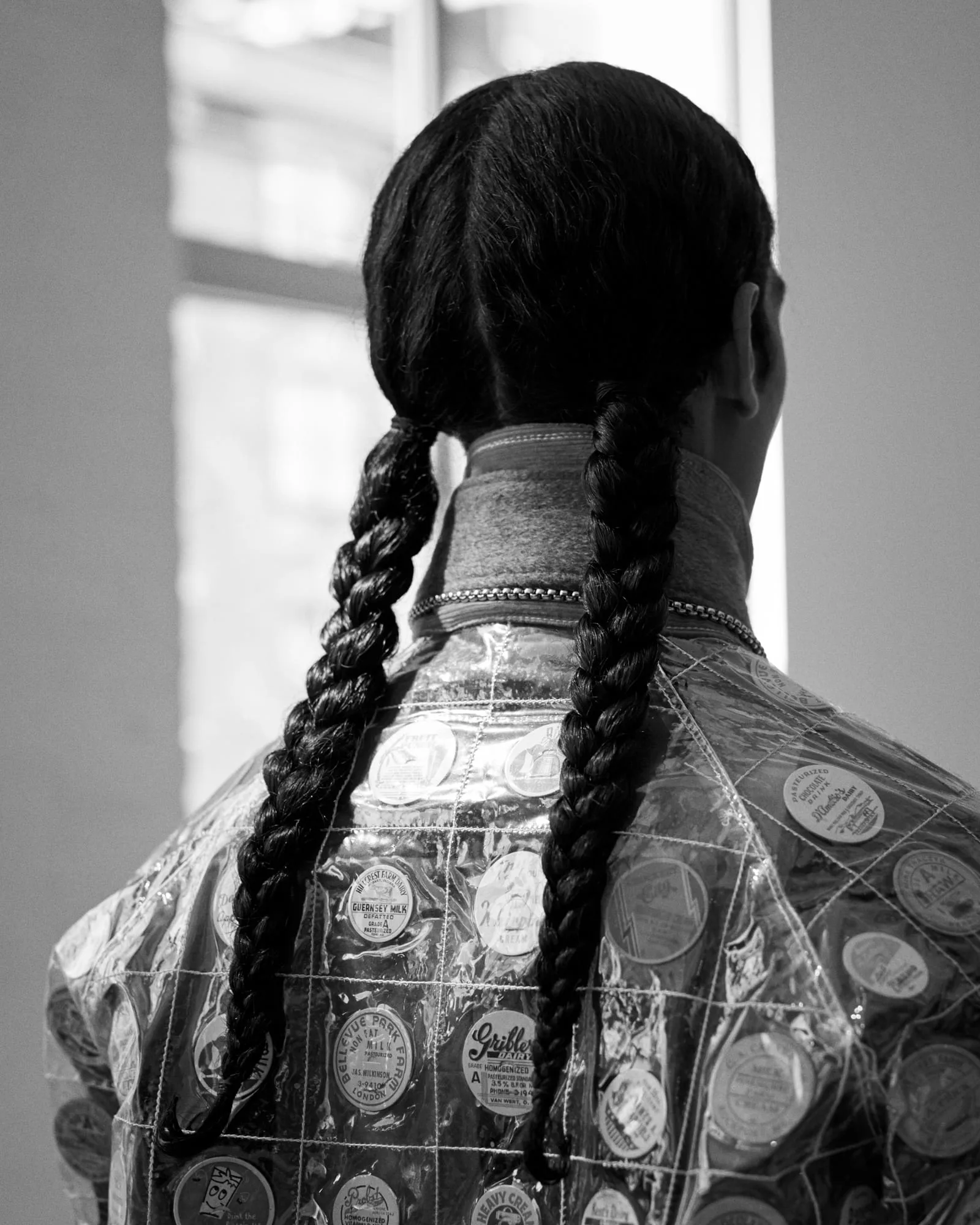 Black-and-white back view of a model with long braids wearing a clear vinyl jacket lined with milk caps, for The Last Magazine.