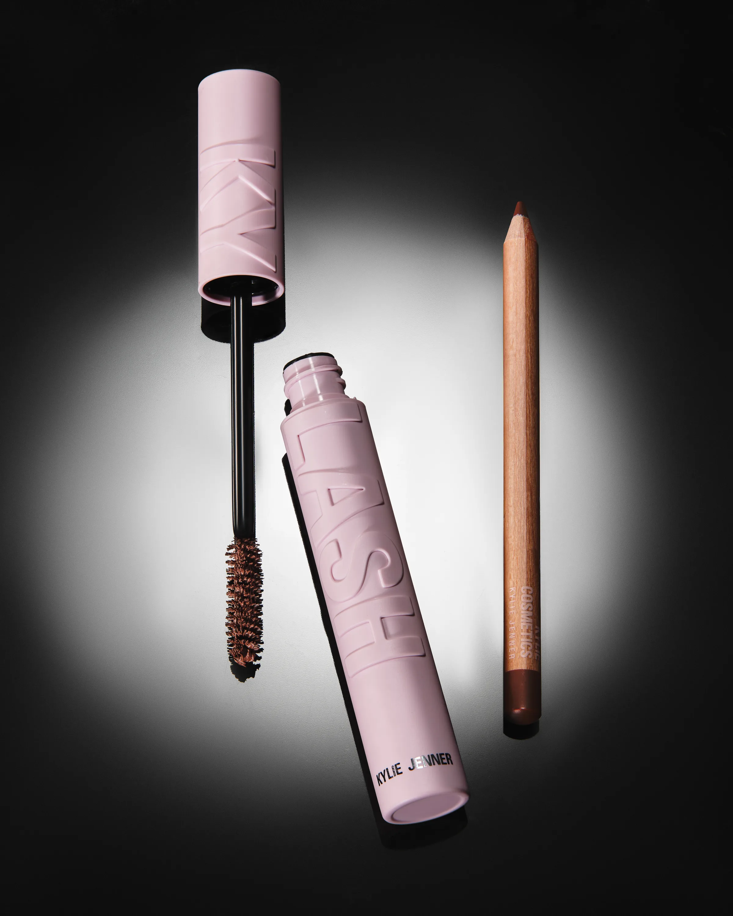 Open pink Kylash mascara with brown wand beside a brown lip pencil under a spotlight on black, for Kylie Cosmetics.