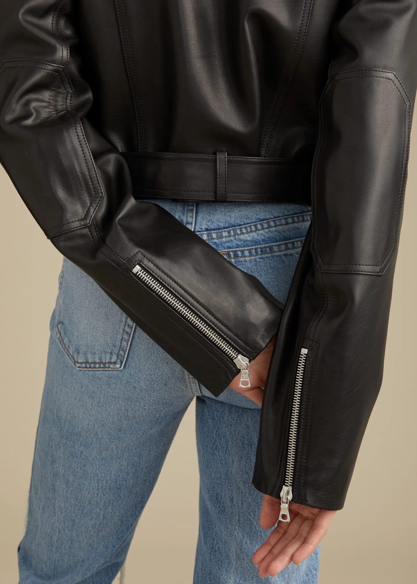 Back view of a black leather moto jacket with zippered cuffs worn over light blue jeans on a beige backdrop, for Khaite.
