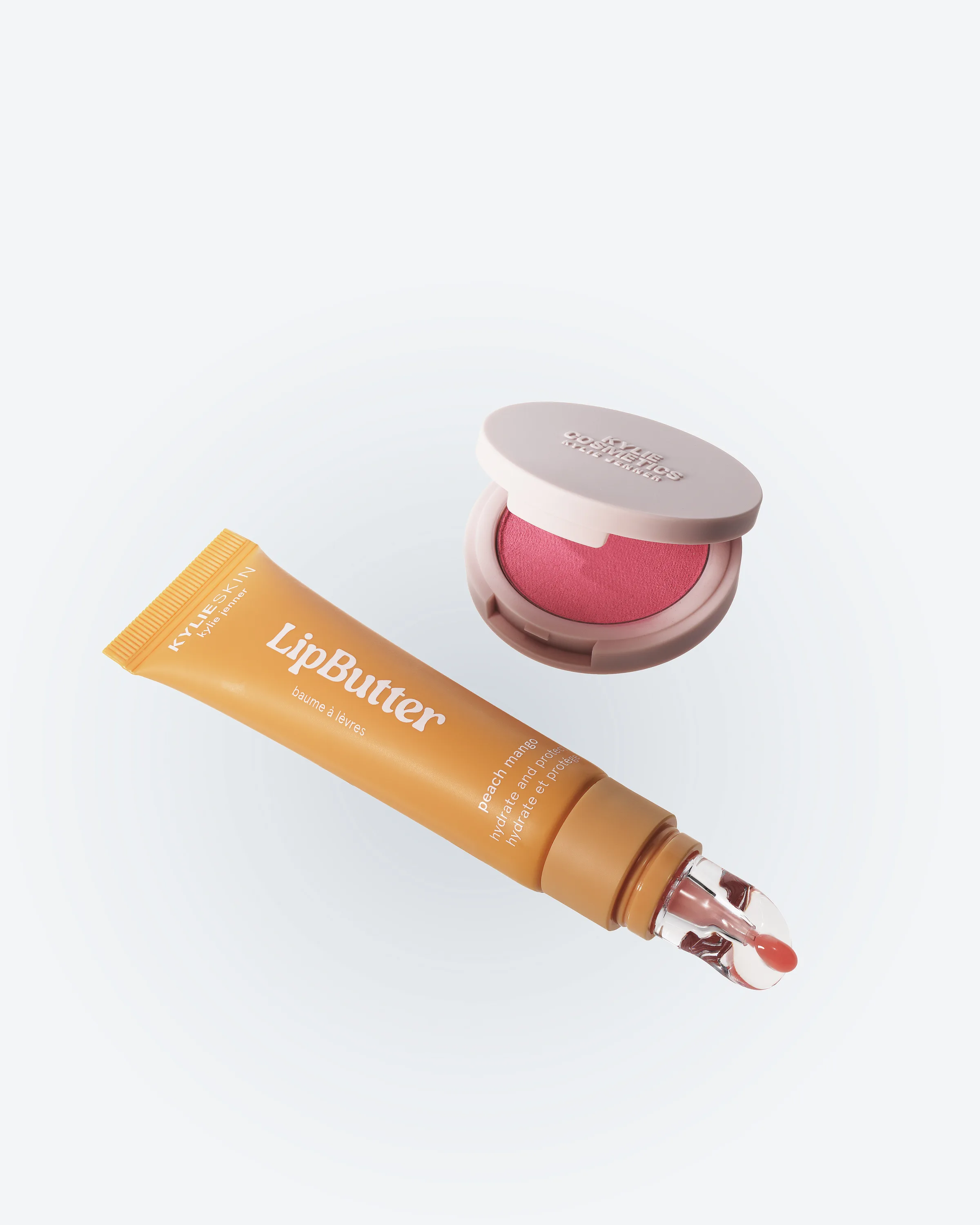 Peach Lip Butter tube floating beside an open pink Cheeky Mood hybrid blush compact on pale grey, for Kylie Cosmetics.