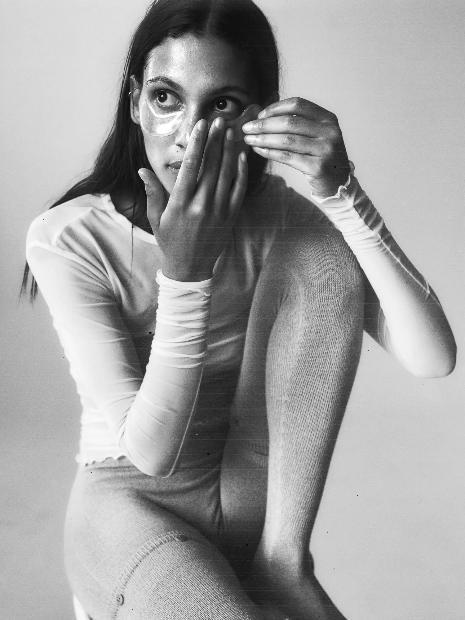 Black and white portrait of a model in a sheer long-sleeve top applying under-eye patches, for The Last Magazine.