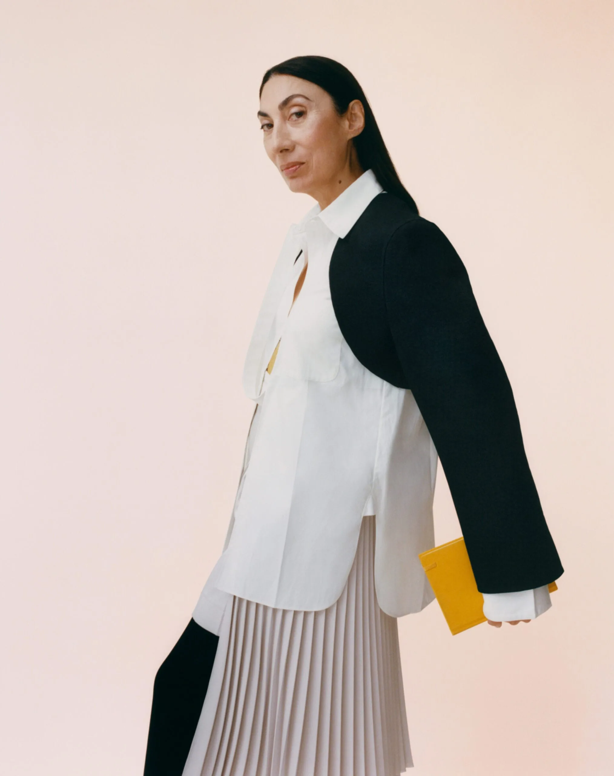 Dark-haired woman in white shirt, black cropped jacket and pleated skirt holding a yellow clutch on blush backdrop, for Interview.