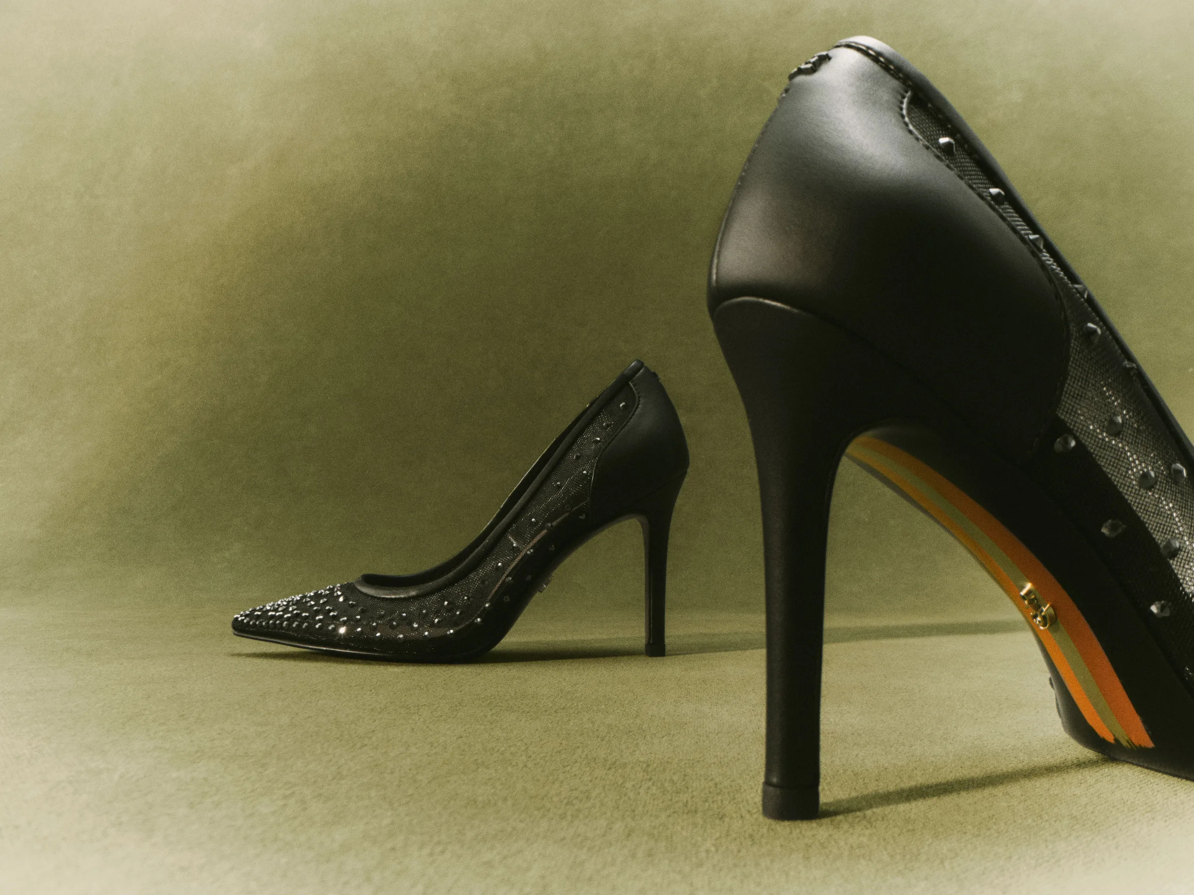 Black studded mesh stiletto pumps posed on an olive green backdrop, one heel in close-up, for Sam Edelman.