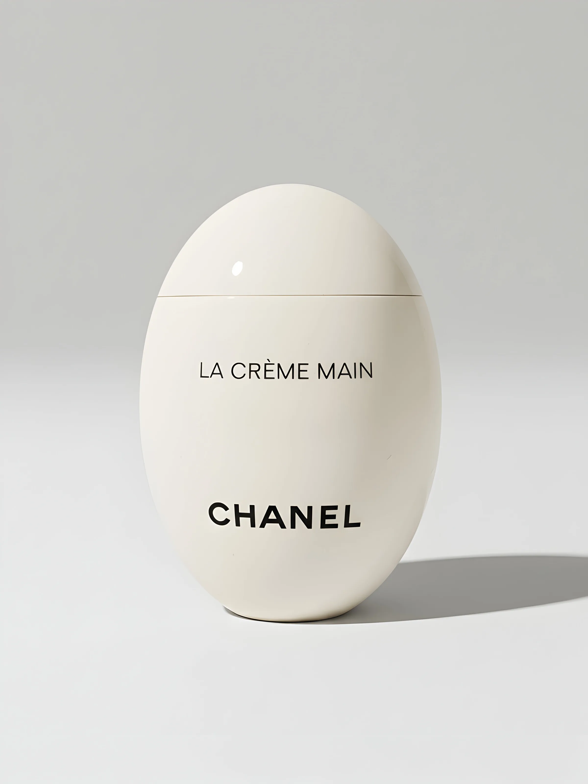 White egg-shaped La Crème Main hand cream bottle on a pale grey background with soft shadow. Chanel AI Concept.