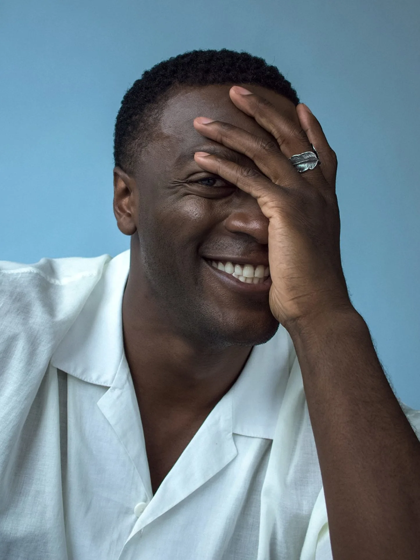 Smiling man in a white linen shirt covering one eye with his hand, wearing a silver leaf ring, on blue, for The Last Magazine.