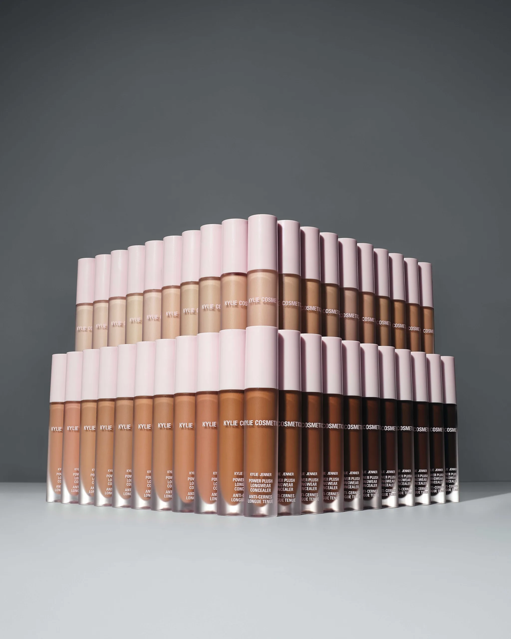 Two tiers of Power Plush concealer tubes in a full range of shades with pink caps on a grey set, for Kylie Cosmetics.