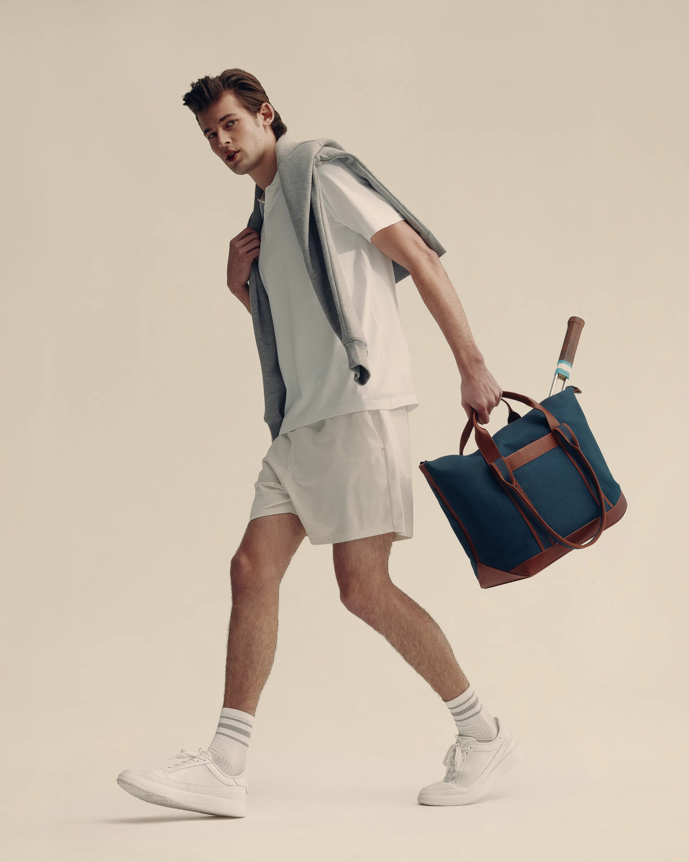 Model walking in white tee, shorts and sneakers, grey sweater on shoulders, carrying a navy tote with racket, for Cole Haan.
