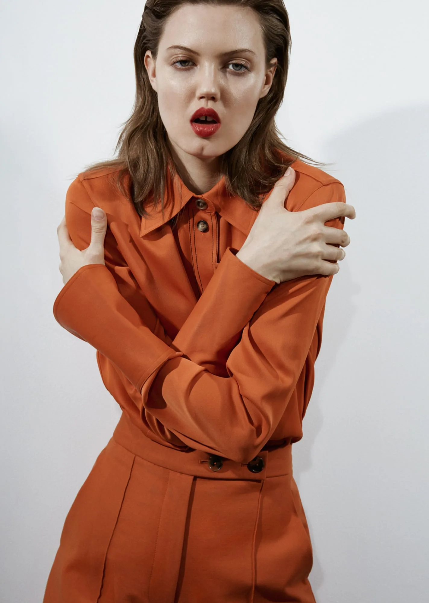 Model in an orange button-front shirt and trousers hugging her shoulders against a white wall, for Puss Puss.