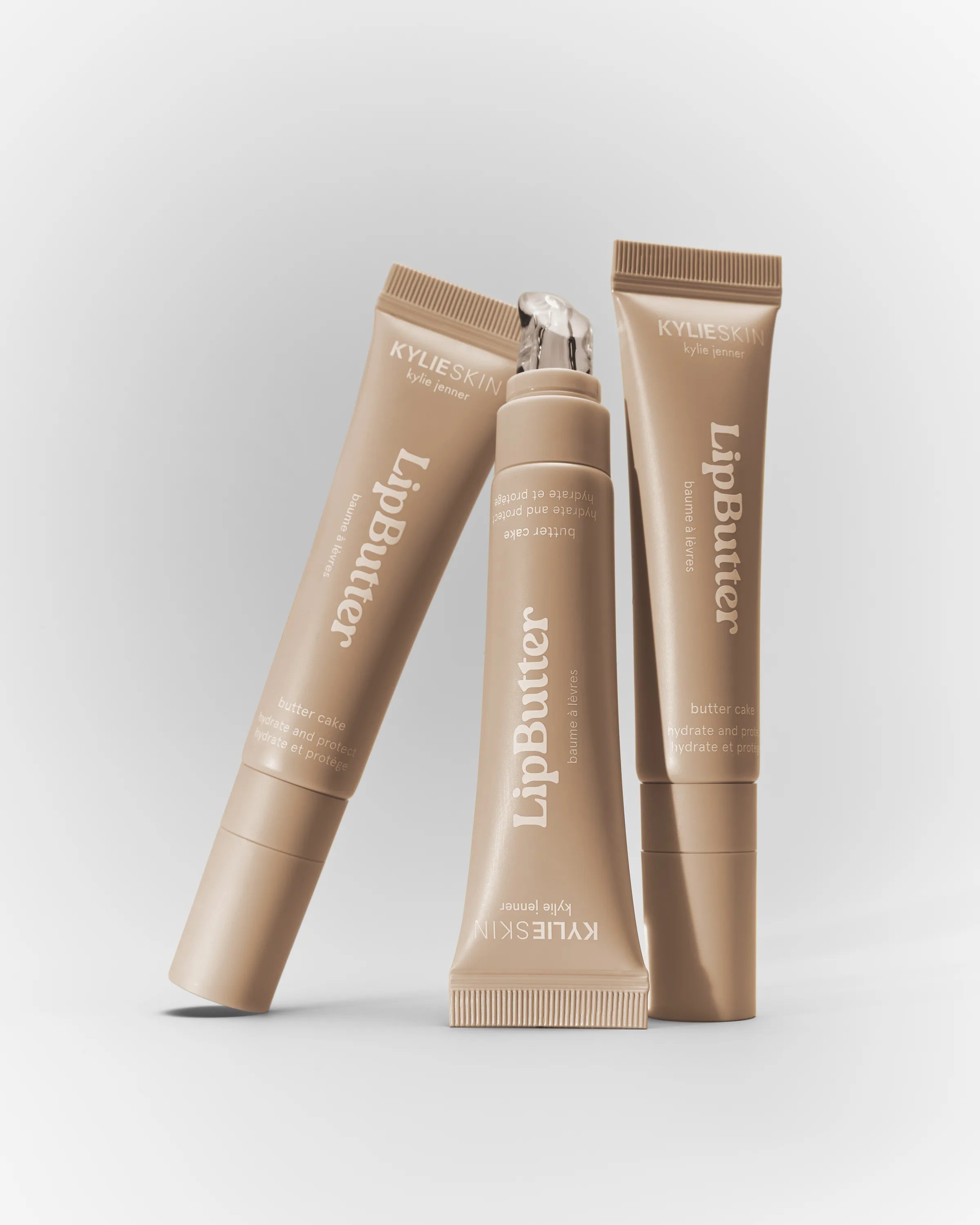 Three beige Kylie Skin Lip Butter tubes in Butter Cake, one standing cap-off, on a pale grey backdrop, for Kylie Cosmetics.