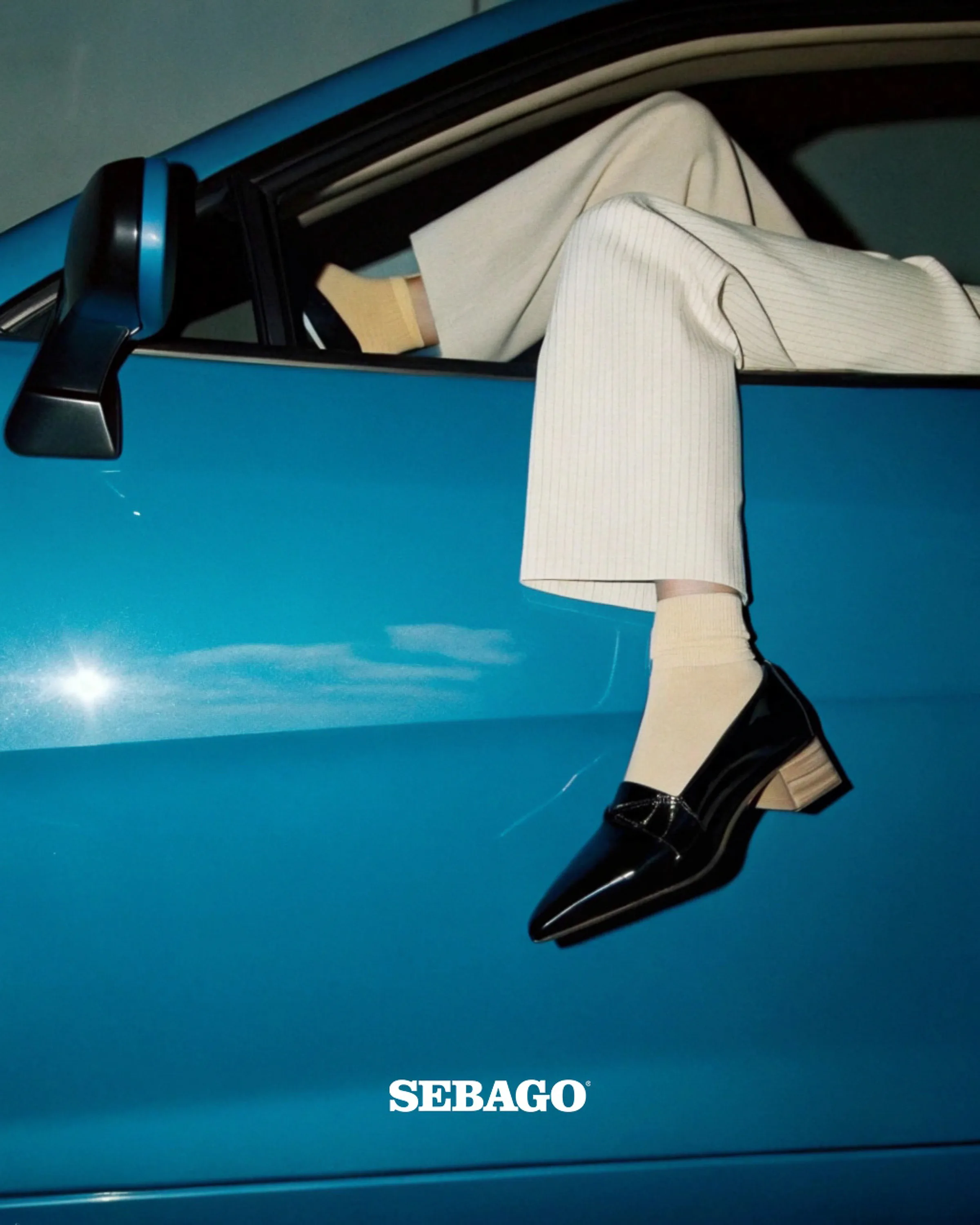 AI image of legs in cream pinstripe trousers and black patent loafers hanging from a blue car window. Sebago AI Concept.