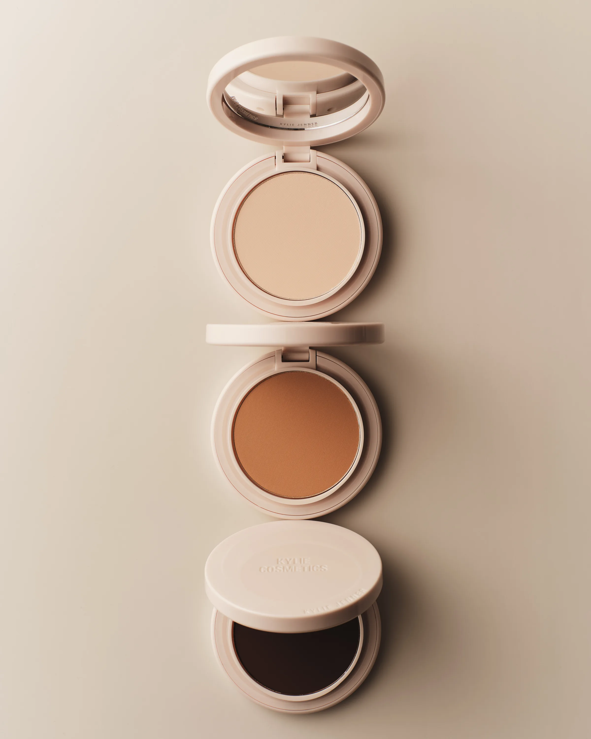 Three open powder foundation compacts in light, medium and deep shades stacked vertically on beige, for Kylie Cosmetics.