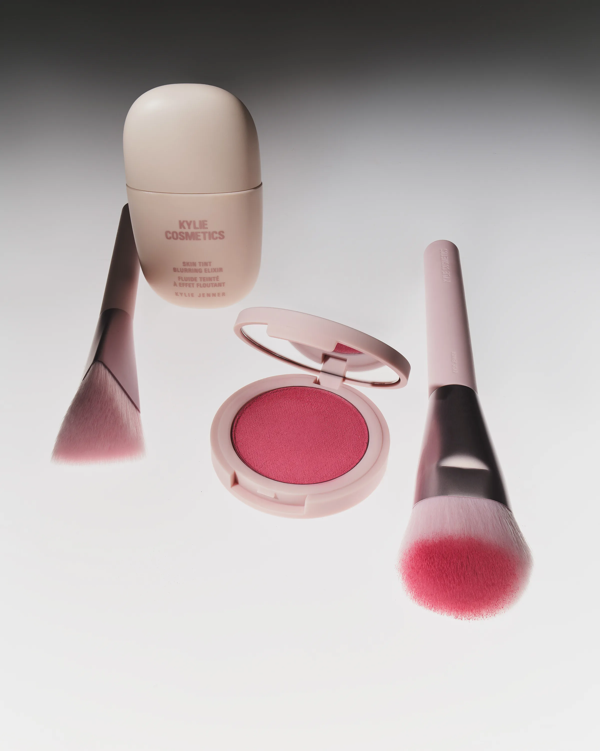 Skin tint bottle, open pink blush compact and two pink makeup brushes on a white surface, for Kylie Cosmetics.