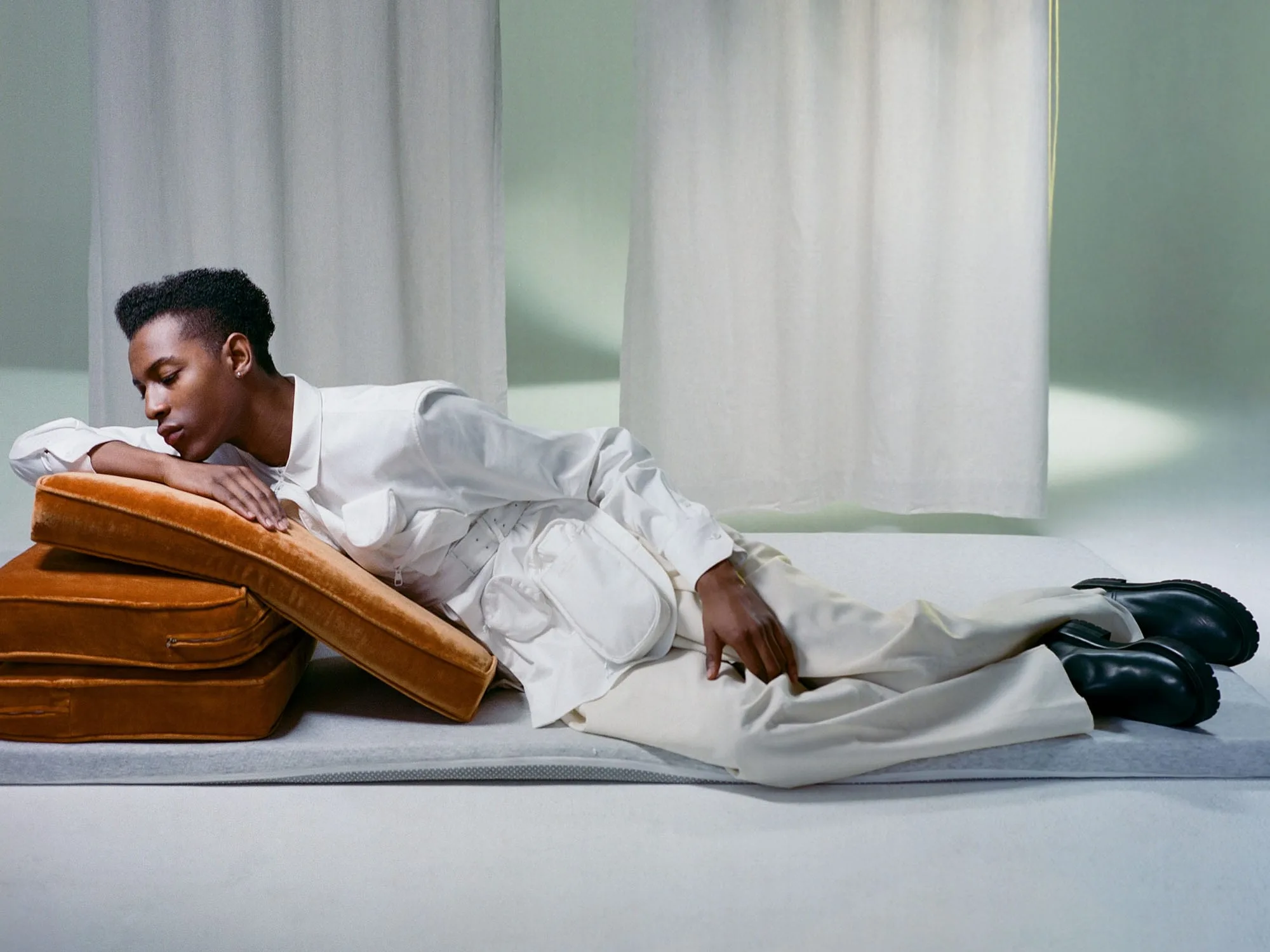 Model in white jacket and trousers reclining on stacked caramel velvet cushions before sheer white drapes, for VMAN.