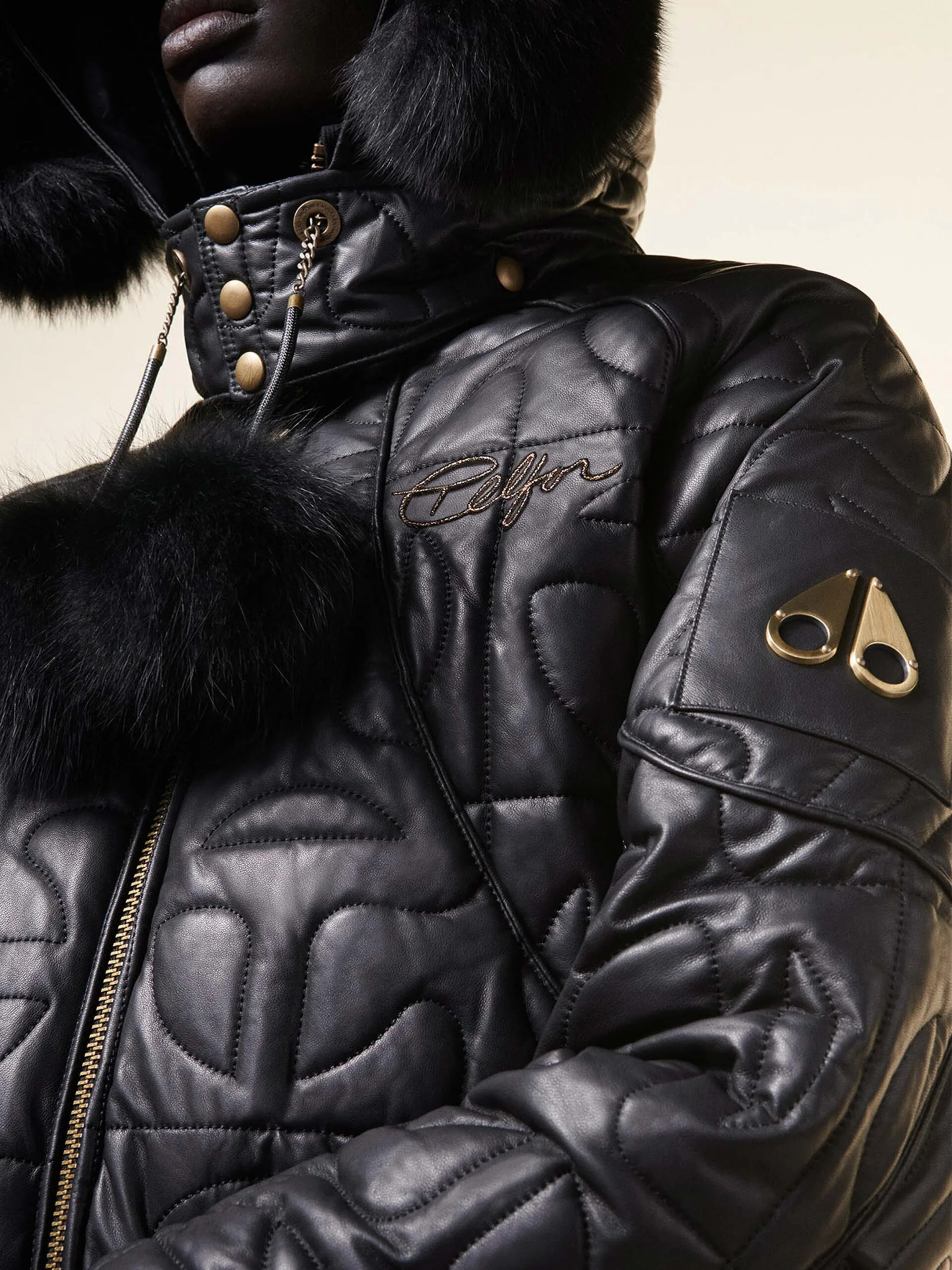 Close-up of a black quilted leather puffer jacket with fur-trimmed hood and gold hardware, for Moose Knuckles.