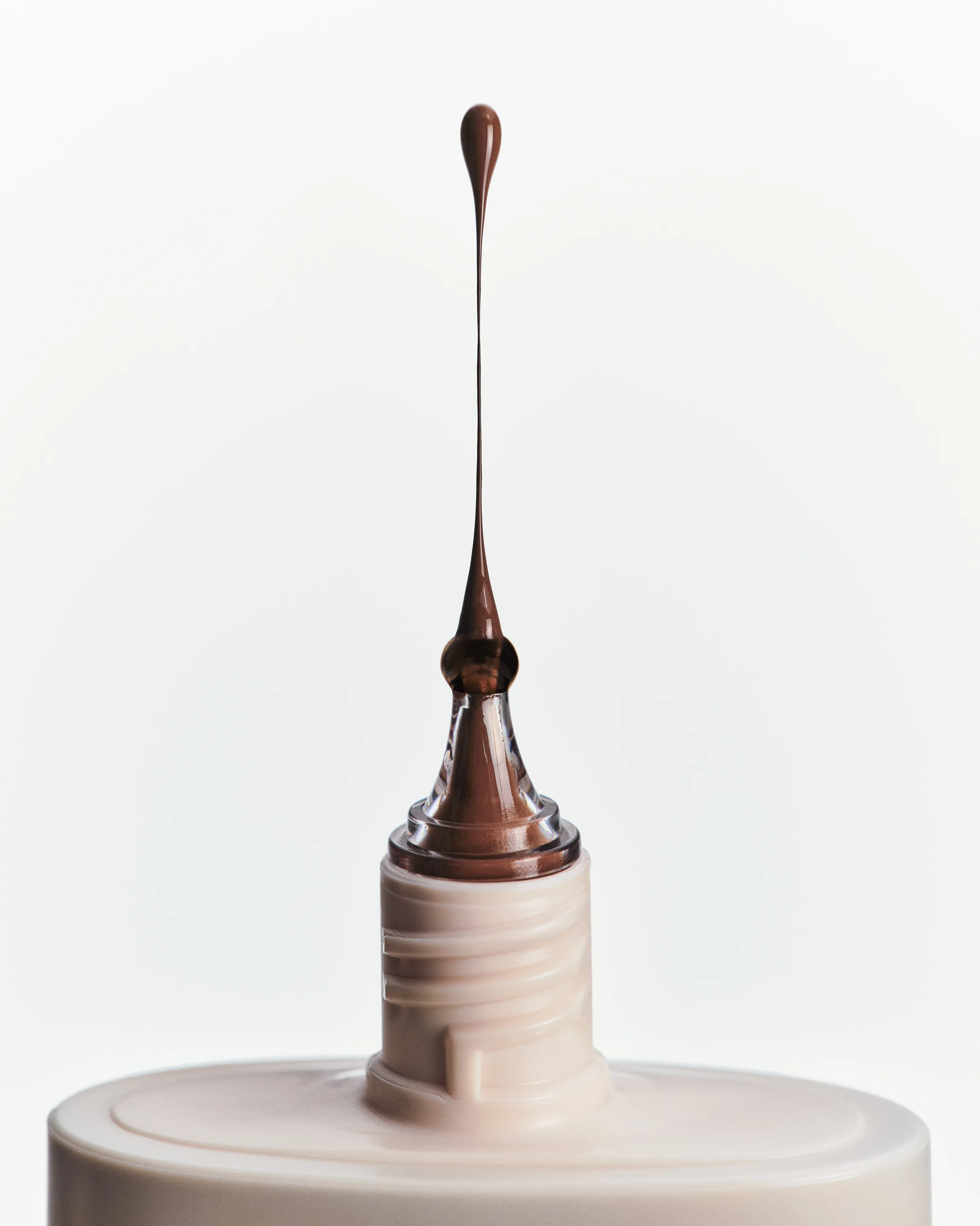 Droplet of brown skin tint rising from a cream bottle nozzle against a white background, for Kylie Cosmetics.