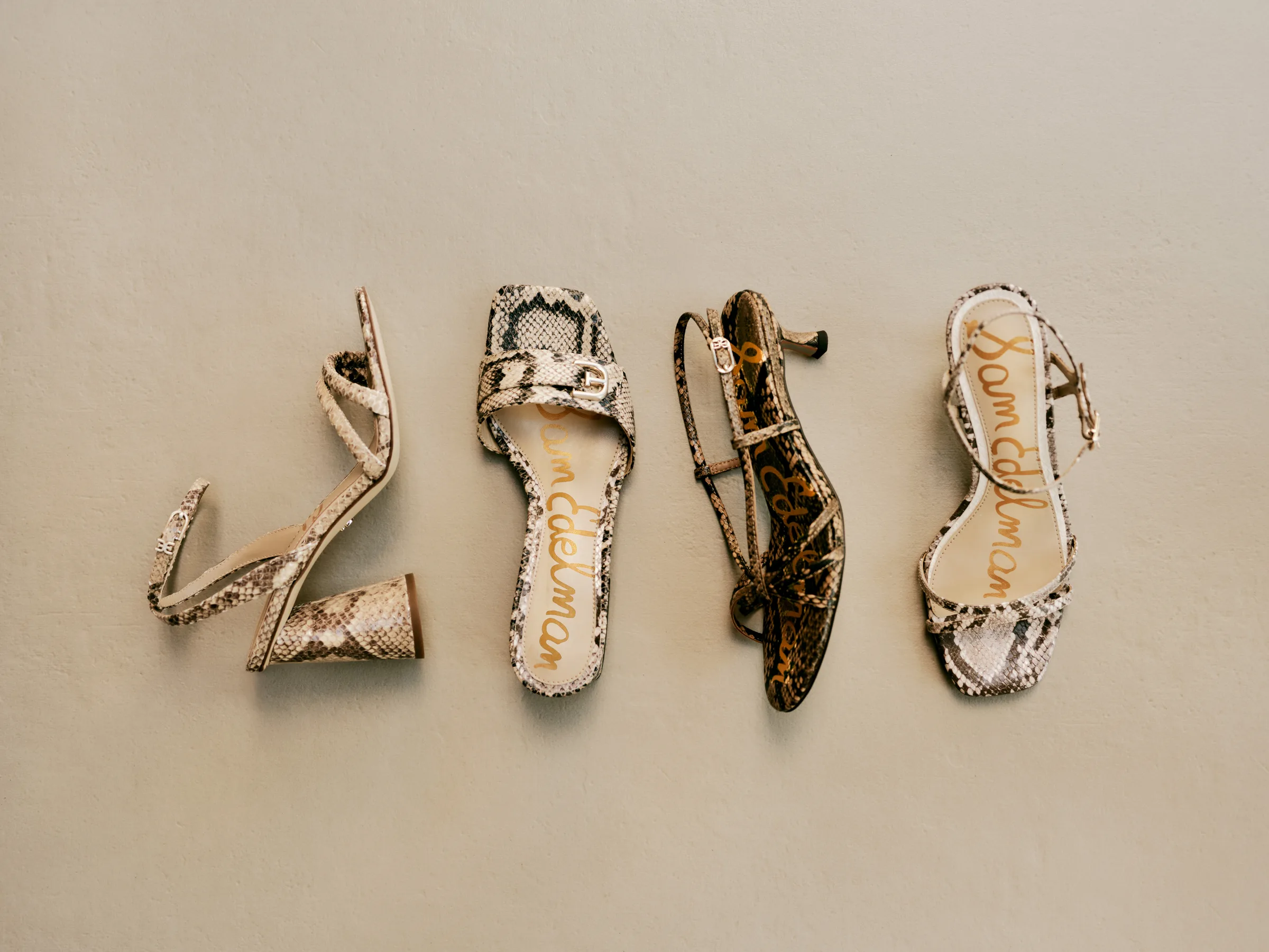 Four snakeskin-print heeled sandals and slides laid in a row on a beige backdrop, for Sam Edelman.