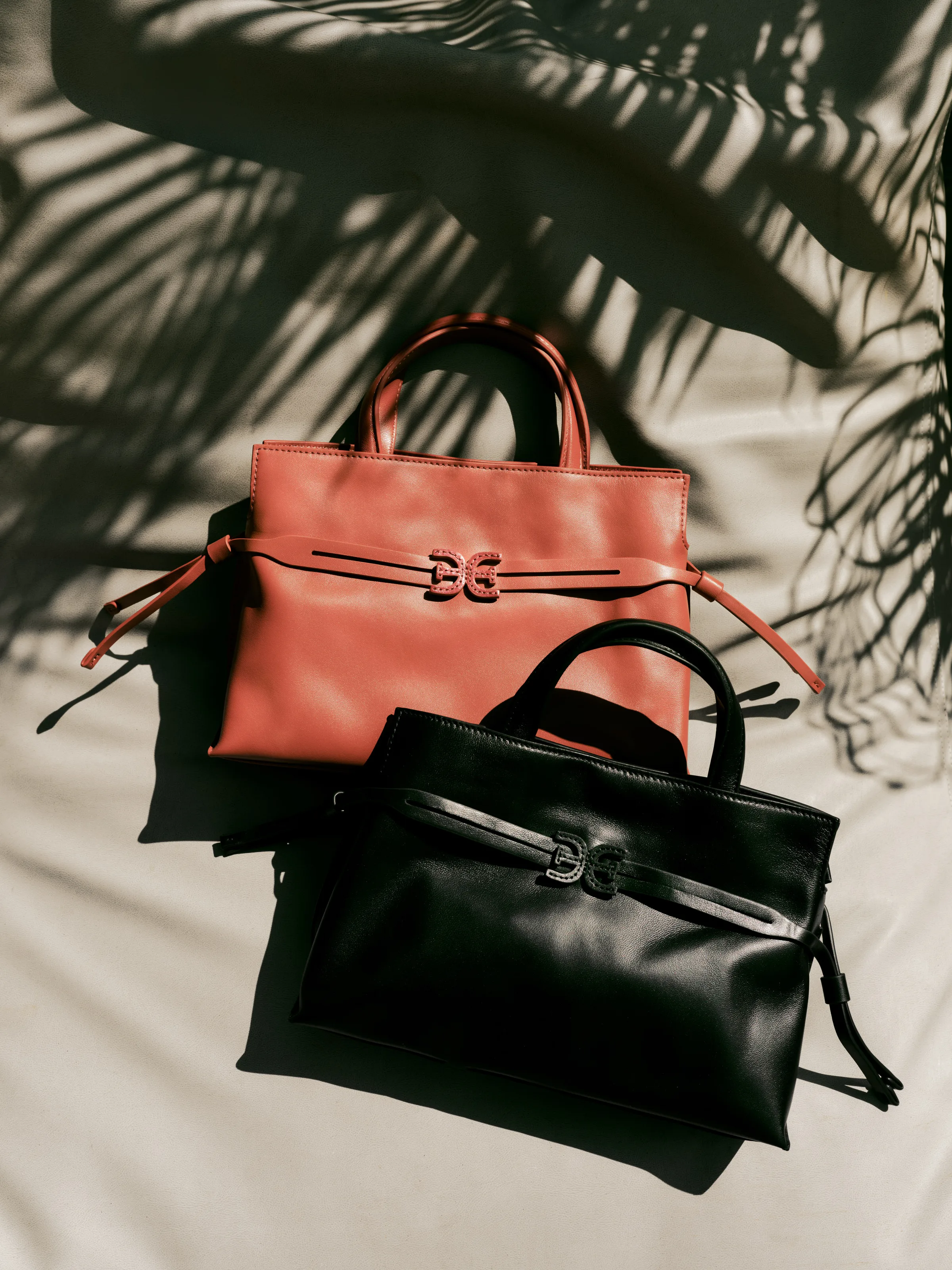 Rust and black leather tote bags with double-E hardware on cream fabric under palm leaf shadows, for Sam Edelman.