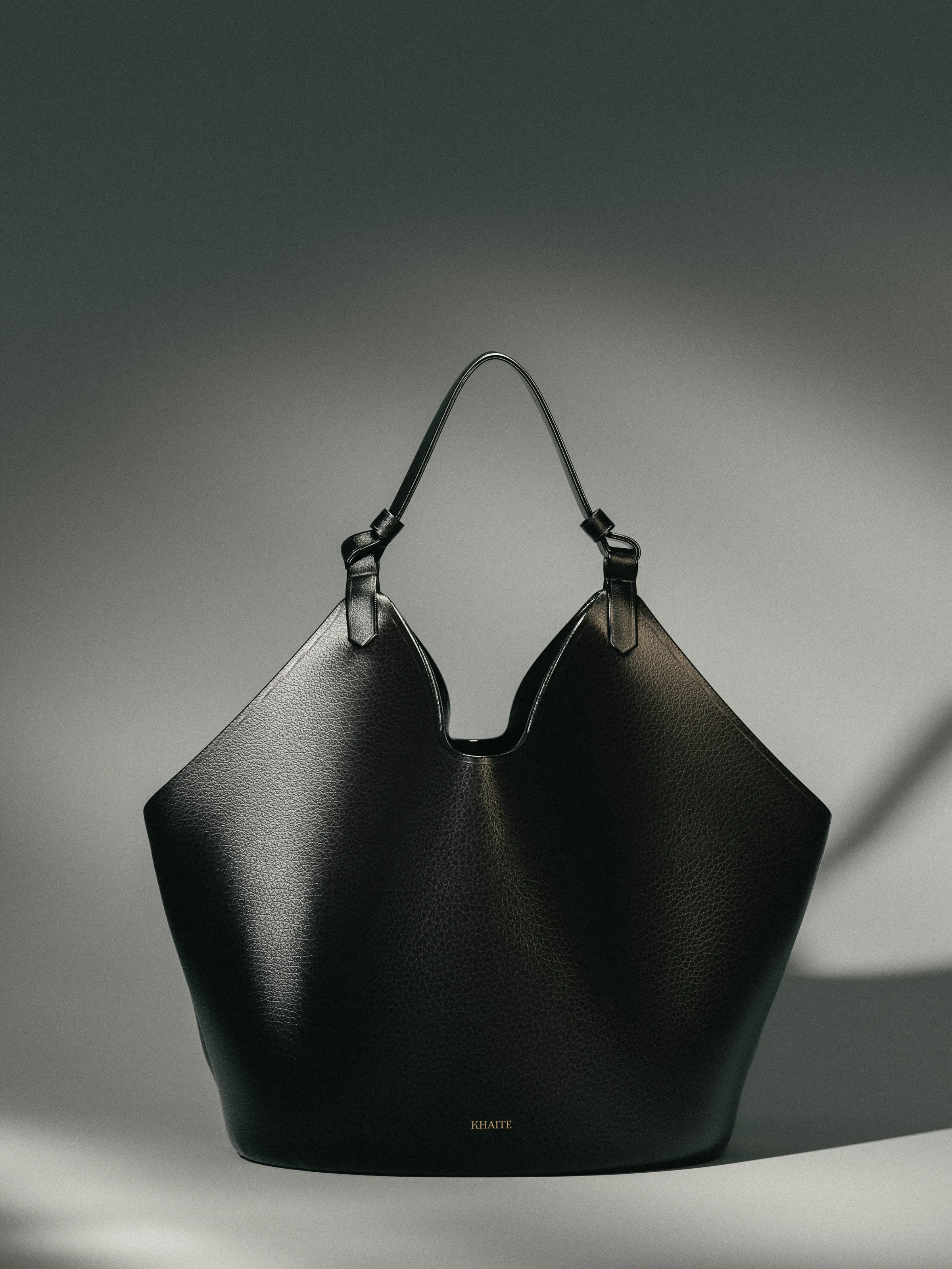 CGI render of a black pebbled leather tote with a knotted top strap under dramatic grey light, for Khaite.