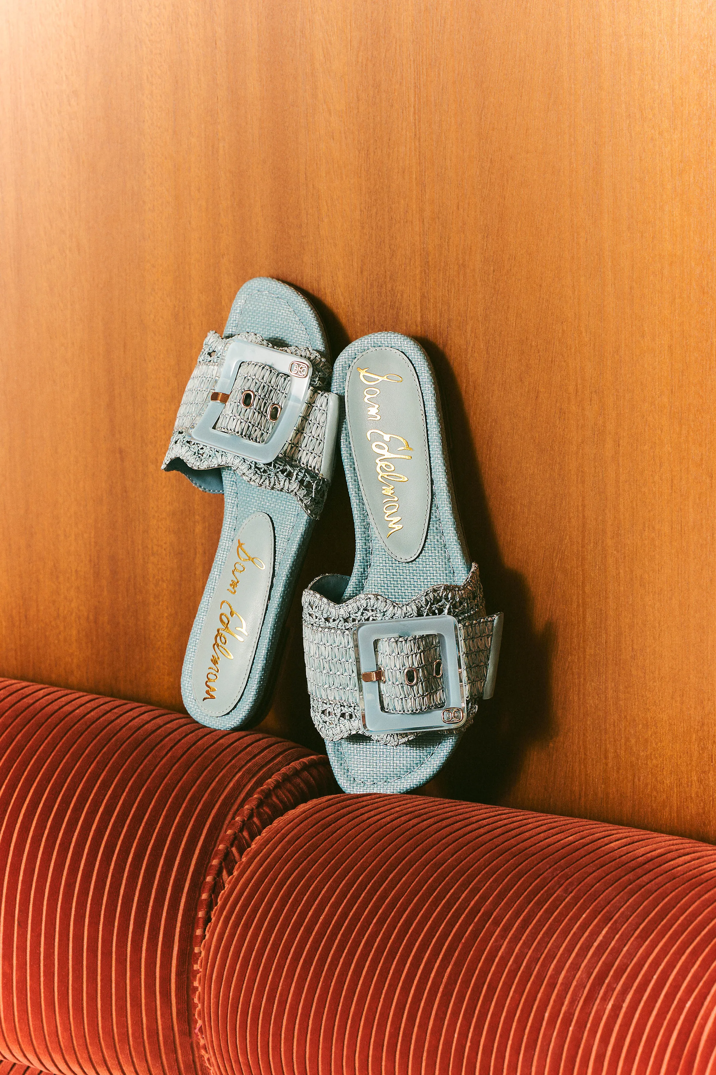 Pale blue woven slide sandals with oversized buckles propped on wood panel above a rust velvet sofa, for Sam Edelman.