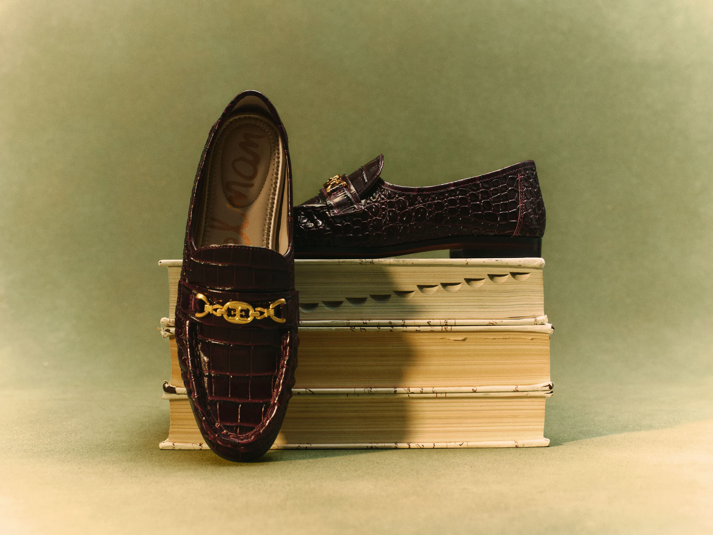 Burgundy croc-embossed loafers with gold chain detail posed on a stack of old books against green backdrop, for Sam Edelman.