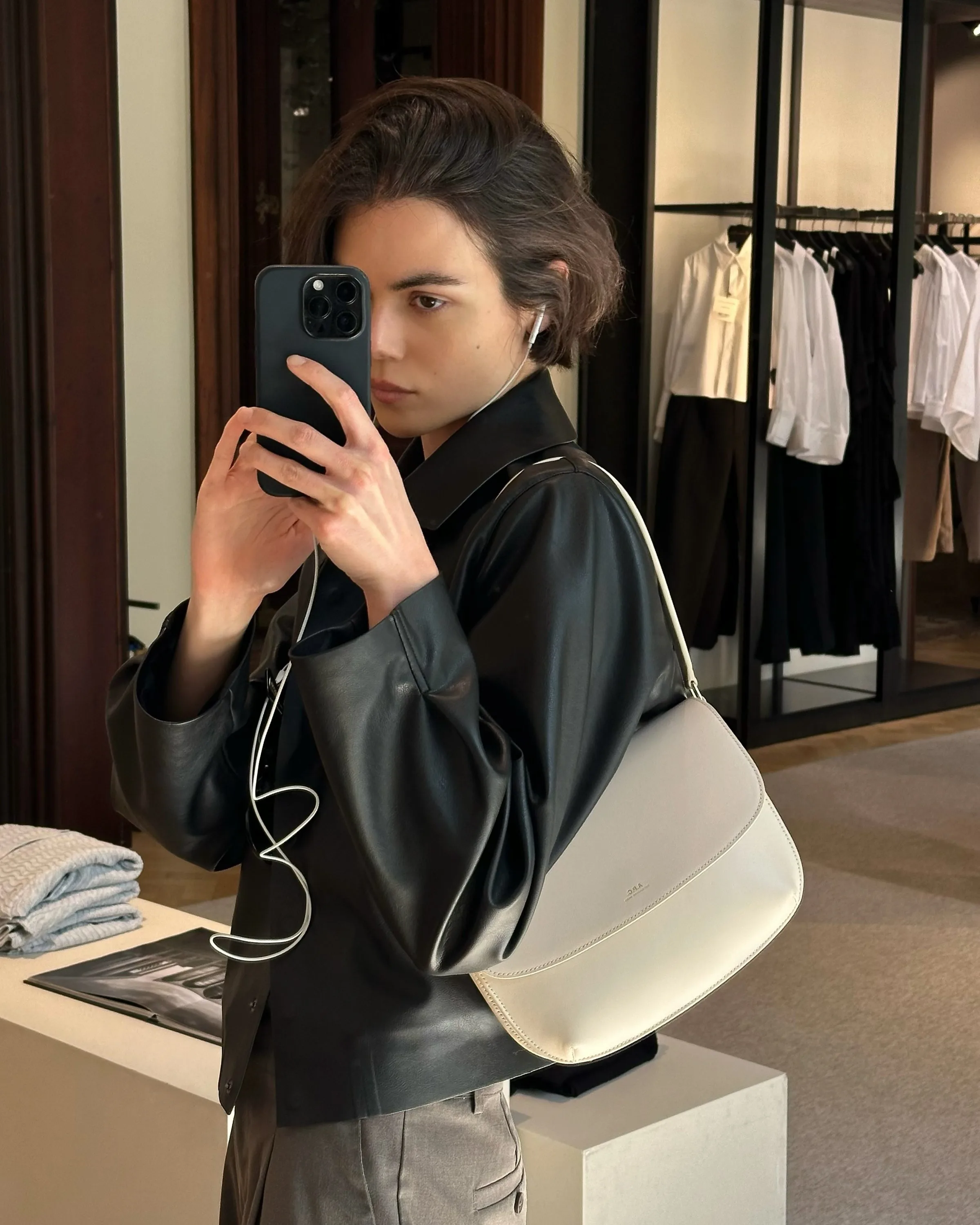 Woman in a black leather jacket takes a mirror selfie in a boutique, wearing a cream shoulder bag and wired earphones.