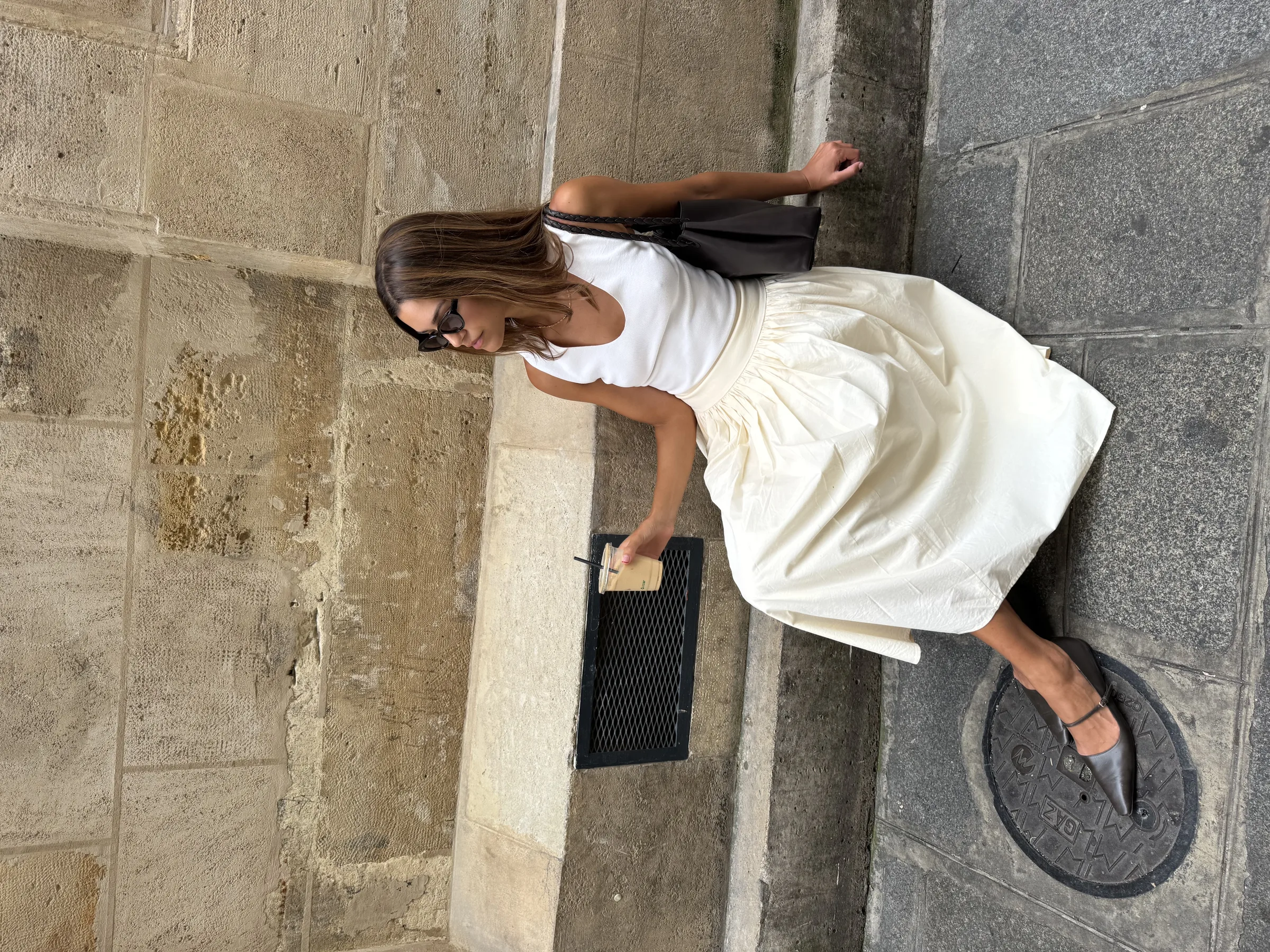 Woman in a white tank, cream midi skirt and black slingback flats sits on a stone ledge holding an iced coffee.