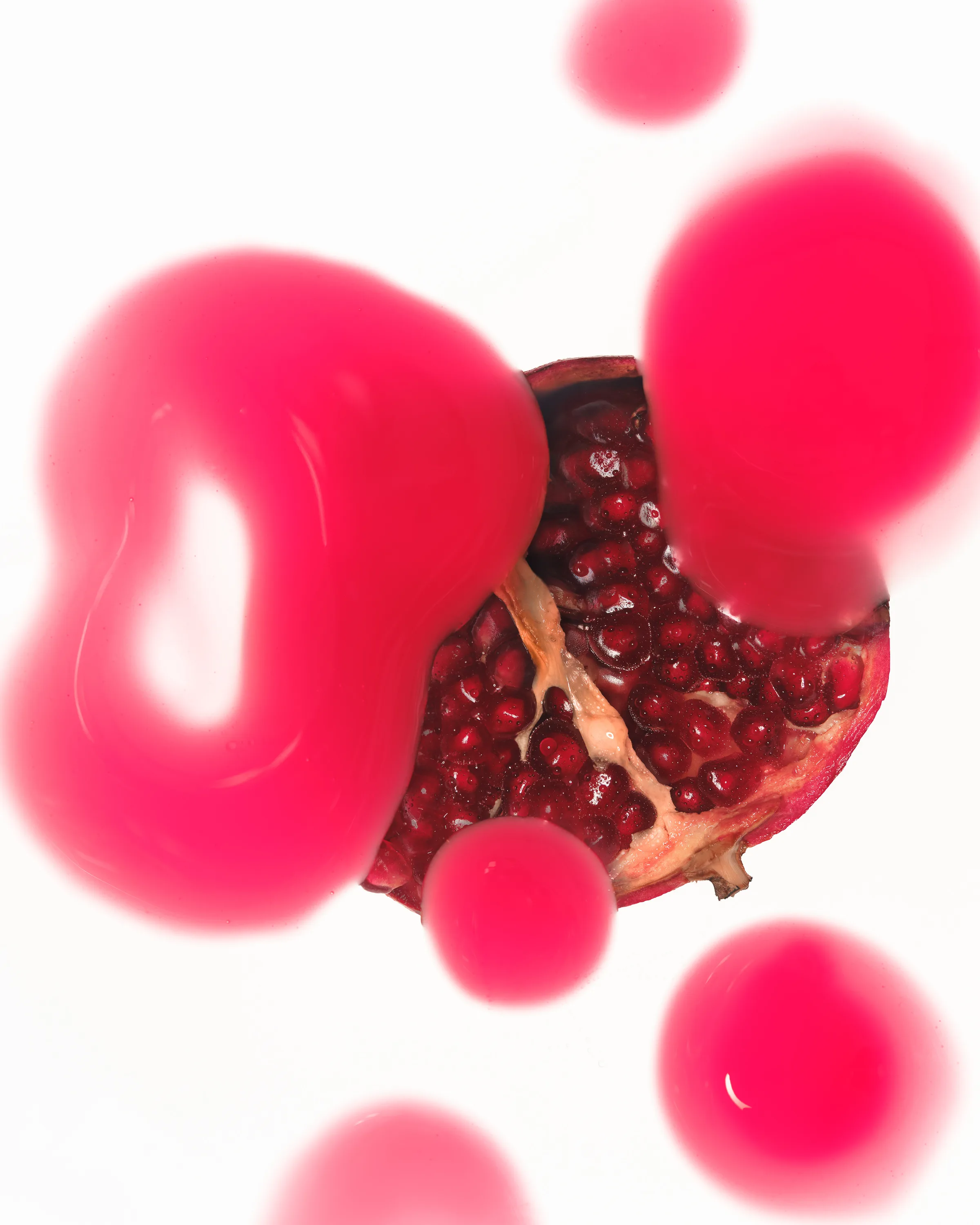 Halved pomegranate seen from above behind glossy pink lip product droplets on white, for Kylie Cosmetics.